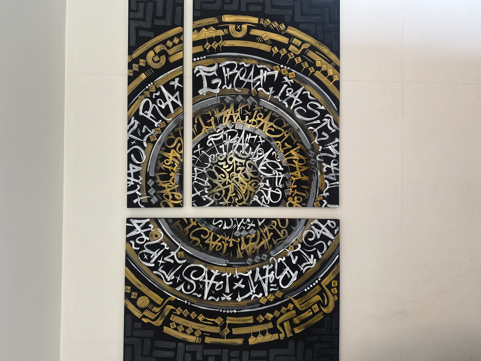The scene captures a multi-panel artwork displayed on a light-colored wall in the city of Madrid, Colombia. The artwork is composed of four distinct panels assembled into a larger rectangular formation, featuring a central design of concentric rings executed in an abstract calligraphic style. The background of the artwork is predominantly black, while the intricate script and decorative elements are rendered in gold, white, and shades of grey. The stylized characters, which do not conform to standard Latin script, form complex patterns that create a mandala-like effect. Small signatures, including 'Julian' and 'ElChico', are visible in the lower left section of the artwork. The wall upon which the artwork is mounted is a plain, off-white surface, with a horizontal line or seam present in the upper wall area. The environment is an indoor setting with even illumination.