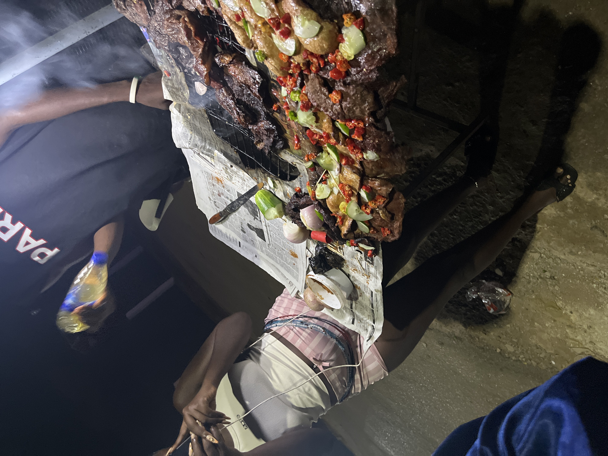 An outdoor nighttime scene in Abuja, Nigeria depicts a group of people gathered around a grill laden with grilled meat. A person in the foreground, wearing a white tank top and pink plaid shorts, is holding a mobile phone. To their right, another person, wearing a black t-shirt with "PAR..." visible on the back, is holding a clear bottle with yellow liquid. The grilled meat, seasoned with red peppers and cucumber slices, is arranged on a metal rack over newspaper. The ground appears to be dirt.