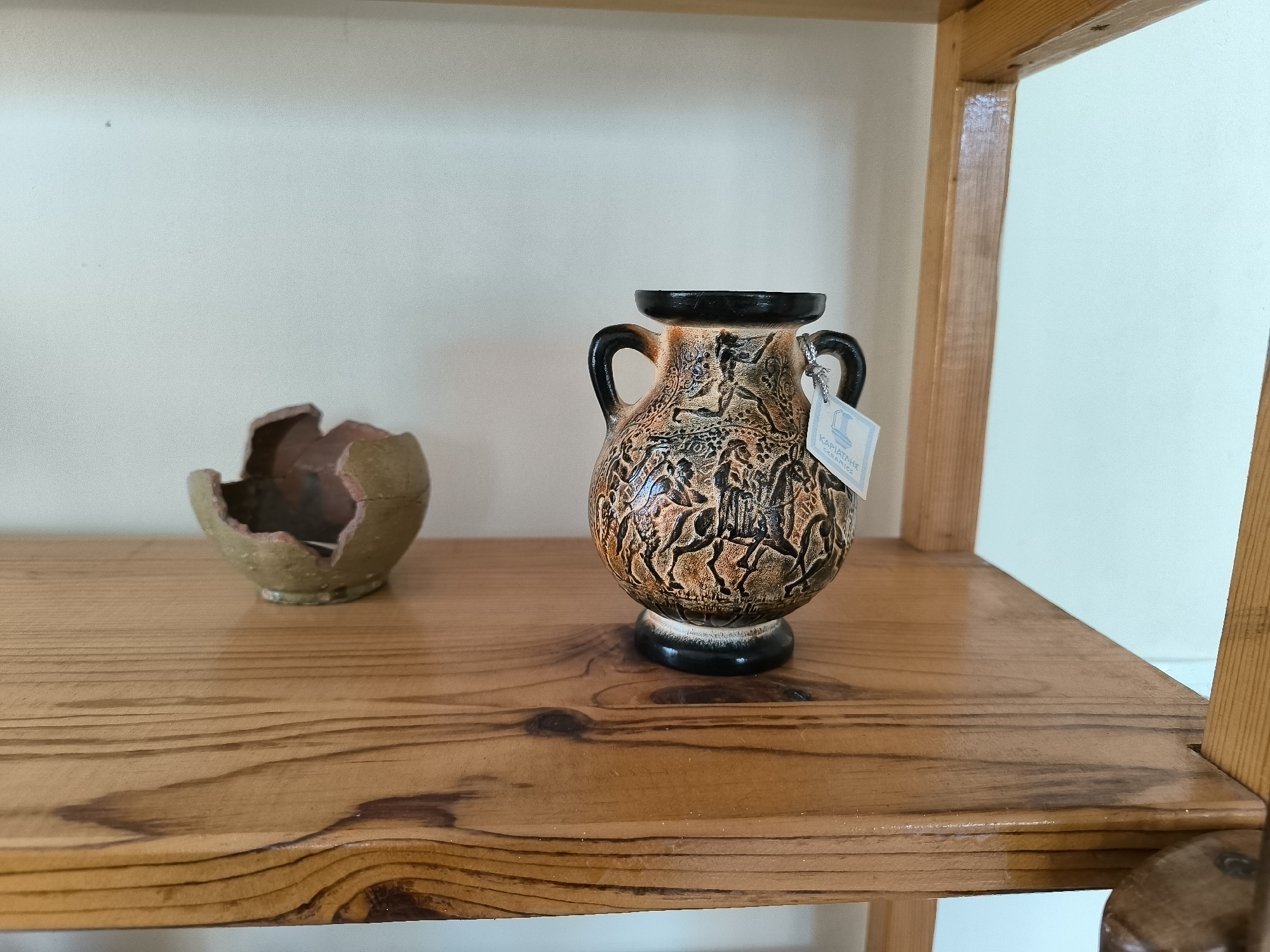 The image shows two ceramic artifacts on a wooden shelf. The artifact on the left is a broken, light green bowl with a reddish interior. The artifact on the right is a dark brown amphora with black handles and a black rim. The amphora is decorated with an intricate pattern of figures in brown and black. A white tag with blue lettering is attached to one of the amphora's handles, reading "KARIATANE CERAMICS". The shelf is made of light-colored wood with visible grain and knots. The background is a plain, off-white wall. The scene is set in the city of Garmen, Bulgaria.
