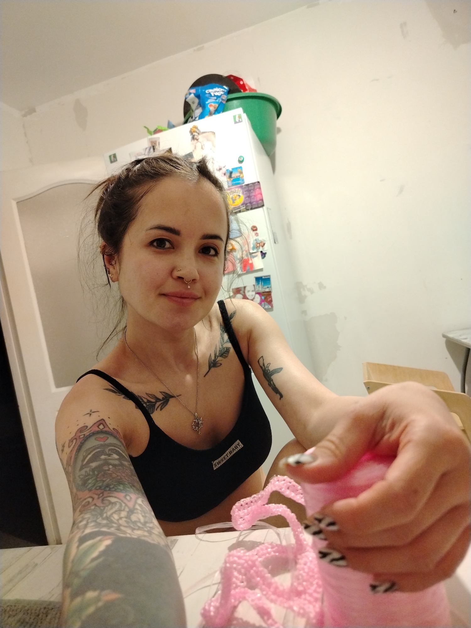 A fair-skinned woman with dark hair pulled back is holding a ball of pink yarn in her right hand. She wears a black tank top and a silver necklace. Her left arm is covered in tattoos. She has a nose ring. In the background, a white refrigerator is visible, covered in magnets and artwork. A green bucket is on top of the refrigerator. A white doorframe is to the left of the refrigerator. The woman is seated at a table. The location is a room with white walls. The location is not specified as Noviy, Russia.