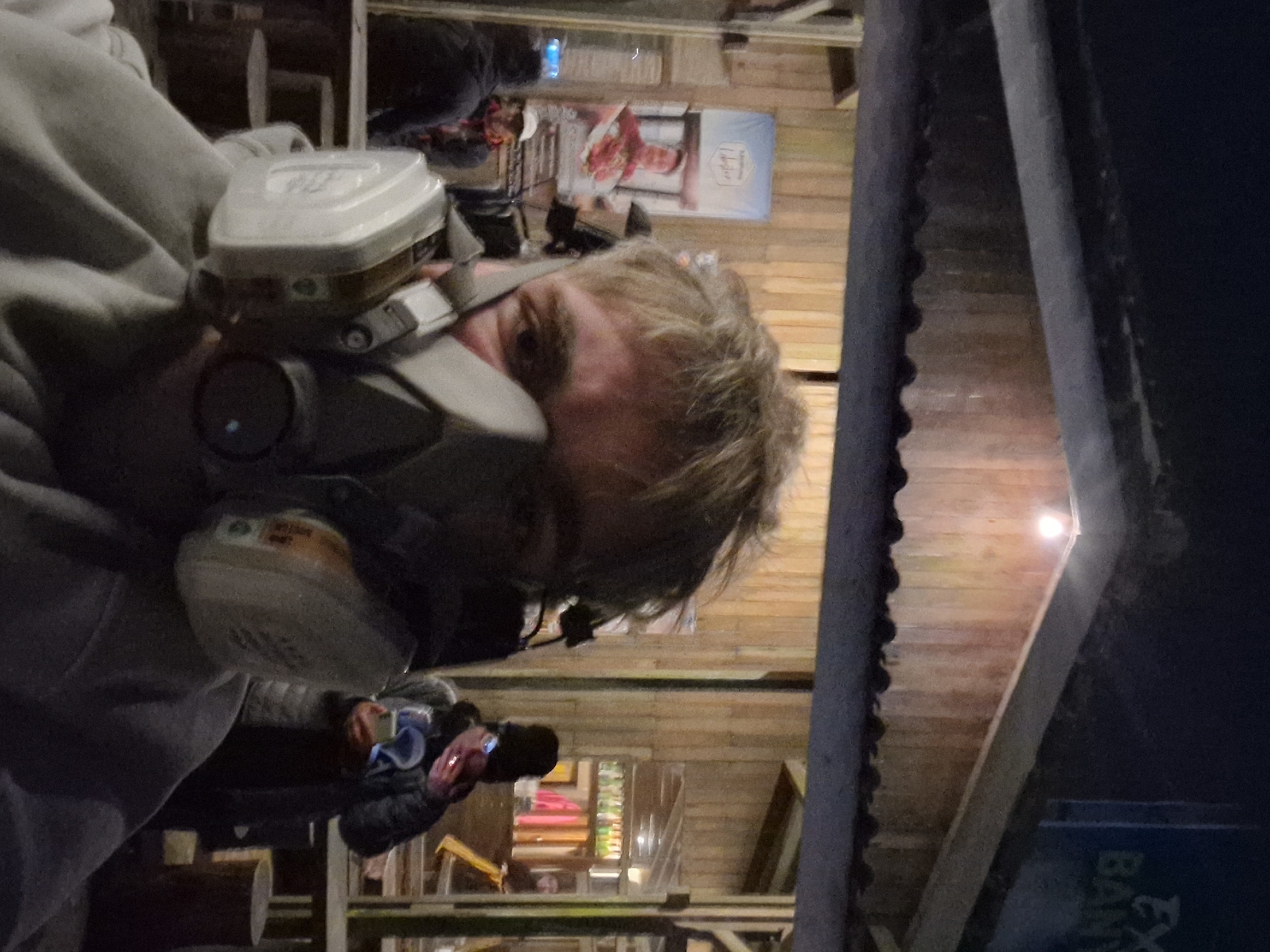 A man wearing a grey hooded sweatshirt and a respirator-type face mask is positioned in the foreground, partially obscuring the view. The background reveals what appears to be an indoor market or event space with wooden structures and displays. A person can be seen photographing something in the mid-ground. In the background, there is signage and additional people. The scene is dimly lit. There is a dark column at the right edge. The location is tagged in Unknown, Indonesia.