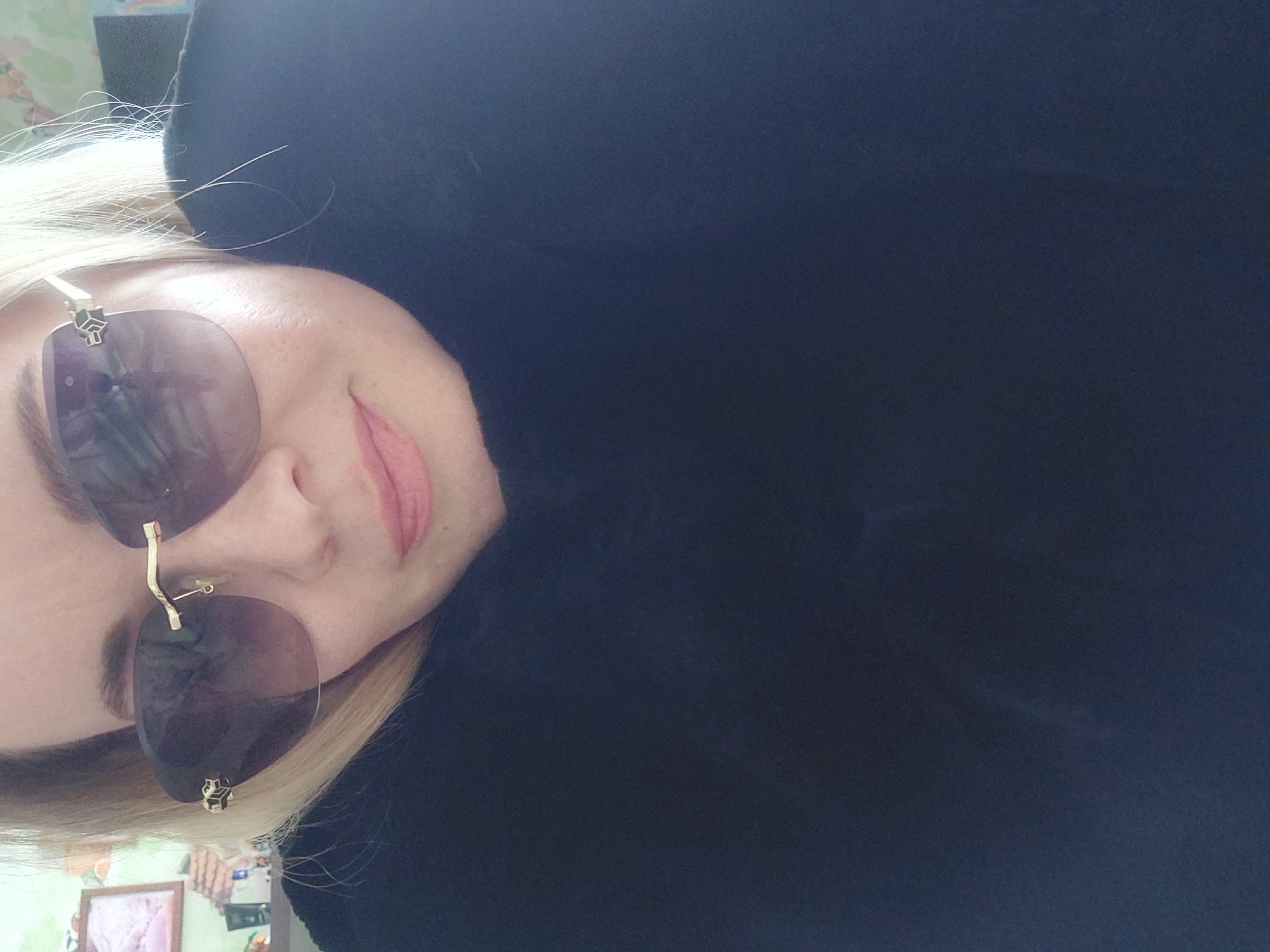 A close-up portrait captures a person, likely female, with blonde hair, tilted to the side and looking towards the viewer. The subject has fair skin and is wearing large, dark-tinted sunglasses with unique gold-colored frames. One arm of the sunglasses is a distinct twisted gold bar, and small decorative elements, possibly star-shaped, are visible on the lenses. A subtle smile is present on the person's lips, which are adorned with pink lipstick.
The person is wearing a dark, possibly black, garment that covers the neck and shoulders. Their blonde hair appears straight, with a few wisps visible near the forehead. The background is divided; the larger portion to the right (when upright) is a plain, dark blue-black surface, while the smaller section to the left reveals a blurry indoor environment, including a hint of a framed picture on what appears to be a wall or furniture. The lighting suggests an indoor setting, likely during the daytime. The overall impression is that of a casual selfie or portrait. No visible text is present in the image. The scene is set indoors, within Chaikovsky, Russia.