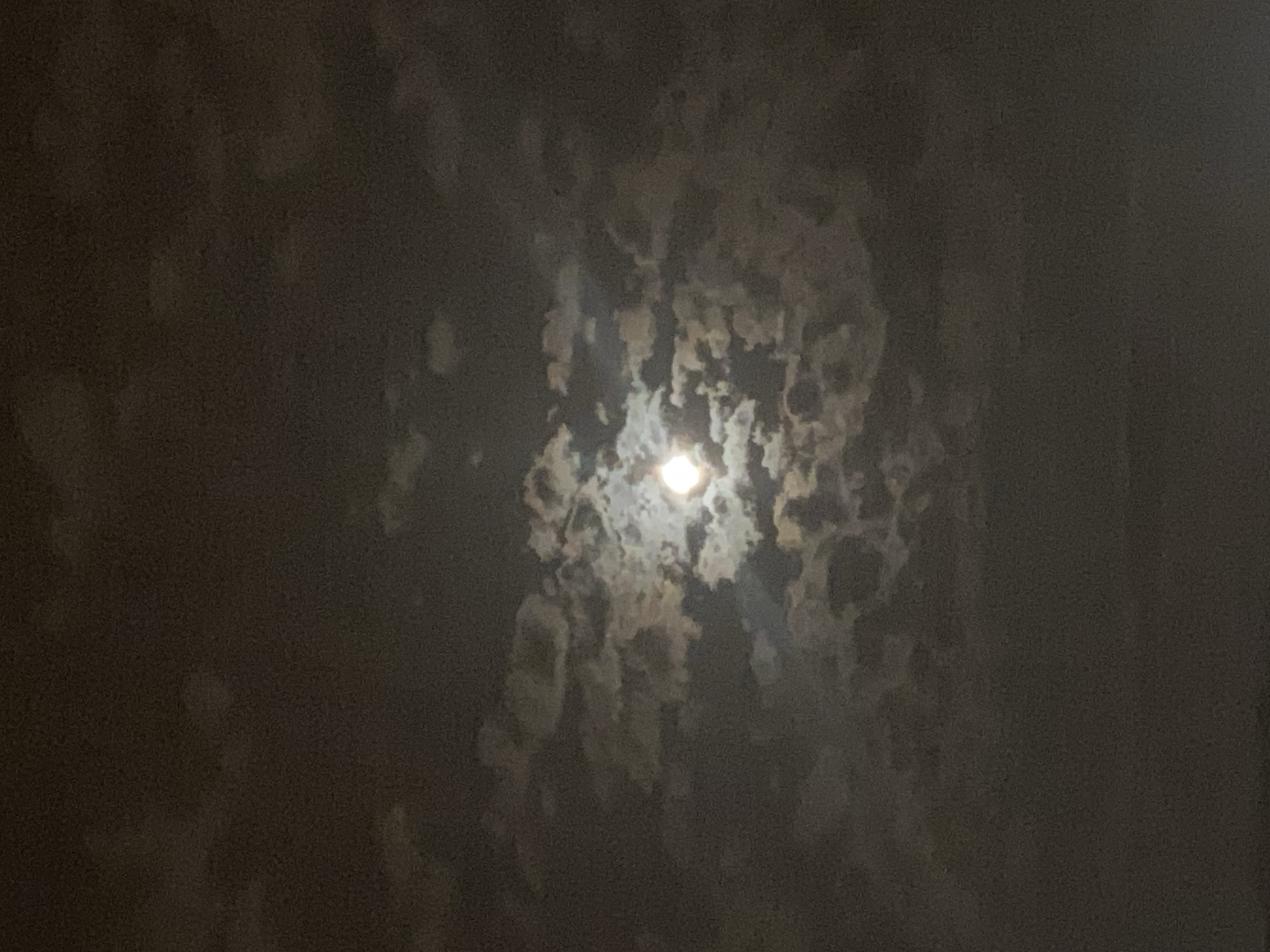 The image is a photograph of the moon partially obscured by clouds, taken at night. The moon is visible as a bright, luminous disc in the center of the frame, with some clouds appearing to glow from its light. The surrounding sky is dark and filled with various cloud formations. The lower portion of the image appears to be a dark, indistinct area, possibly a landscape or body of water. There are no people, objects, or actions clearly discernible in the image. The location is identified as Iași, Romania, though no specific landmarks or urban features are visible to confirm this.