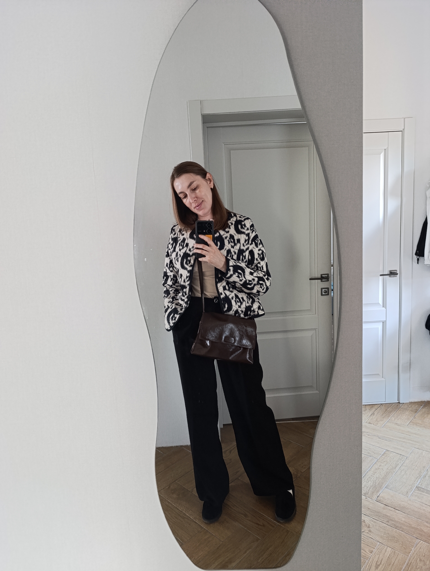A woman stands in front of a mirror, taking a selfie with her smartphone. She is wearing a patterned black and white jacket, a tan top, black pants, and black shoes. A brown leather crossbody bag hangs from her shoulder.
The woman is positioned in what appears to be an indoor setting, likely a hallway or entryway, with a herringbone pattern wood floor. Two white paneled doors are visible in the background, one slightly ajar. Clothing hangs on a rack partially visible through the open door.
The image appears to be a reflection in a distorted or uniquely shaped mirror, creating a slightly warped view of the scene.
While the specific location within Chaikovsky, Russia, cannot be determined solely from the image, it can be stated that the setting is an interior space with specific architectural details and design elements.