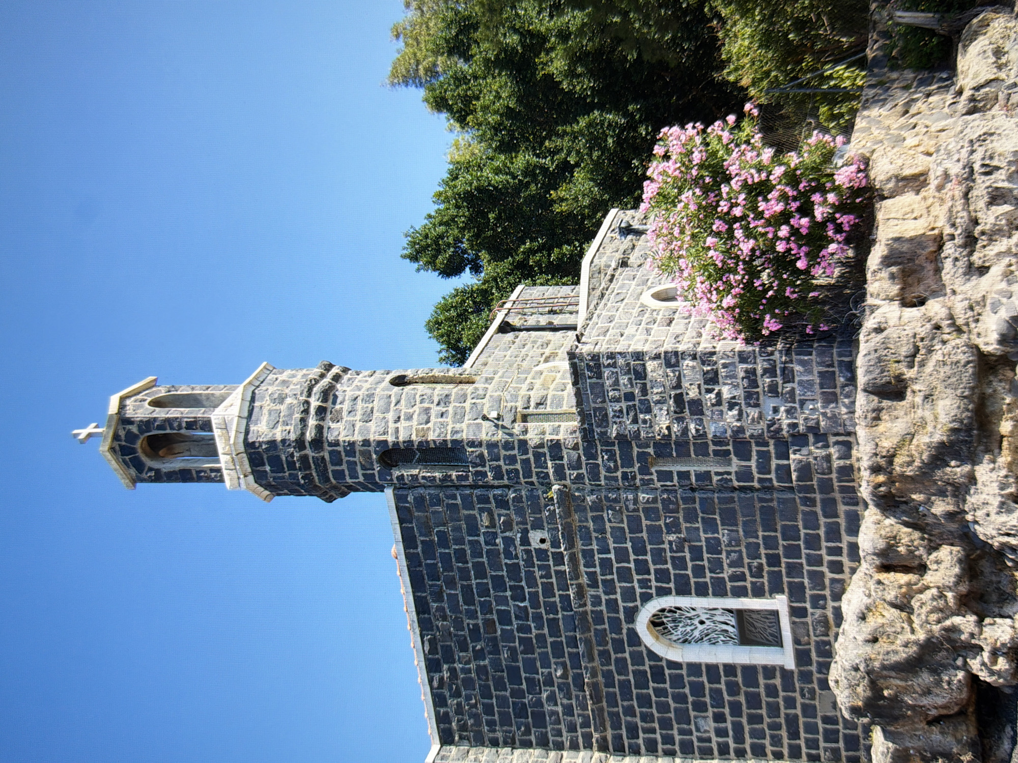 The image shows a stone church with a bell tower and a cross on top, set against a clear blue sky. The church is constructed of dark, rough-hewn stones, with lighter stone accents around the windows and the roofline. The bell tower has arched openings and a conical roof. In front of the church, there is a bush with bright pink flowers, and a rocky, uneven terrain. Tall green trees are visible in the background. The scene appears to be bathed in bright sunlight. This image does not depict the city of Kyiv, Ukraine.