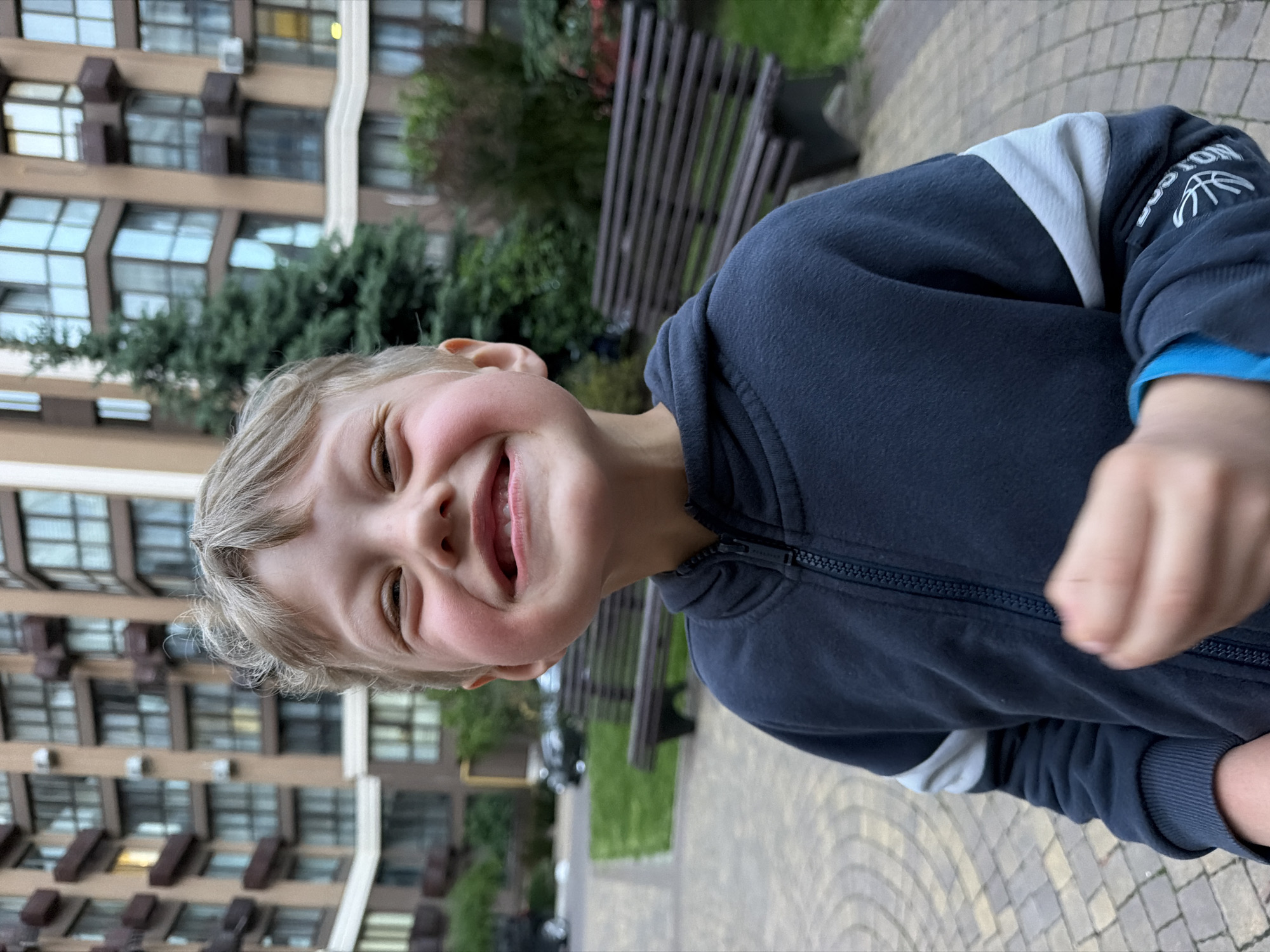 A young boy with light hair and rosy cheeks is captured in a joyous, close-up shot, looking directly at the camera with a wide, open-mouthed smile, his eyes crinkled with happiness. He appears to be between 5 and 8 years old. He is wearing a dark blue zip-up hoodie with a white/light grey stripe or panel on the upper left arm. White text "BOSTON" and a basketball icon are visible on the upper part of the same sleeve. A bright blue layer is visible under the cuff of his right sleeve.
The image is rotated 90 degrees clockwise. The background reveals a modern multi-story residential building with light brown/beige walls and numerous large, dark-framed windows, some with dark rectangular protrusions. In the middle ground, behind the boy, there are dark green bushes or small trees, and a slatted wooden park bench with dark supports. The ground is paved with light-colored, interlocking pavers. The lighting is soft and even, suggesting it is daytime, possibly overcast or late afternoon. The scene takes place in an outdoor residential area, likely a courtyard or park. The location context is Sofiivska Borshchahivka, Ukraine.