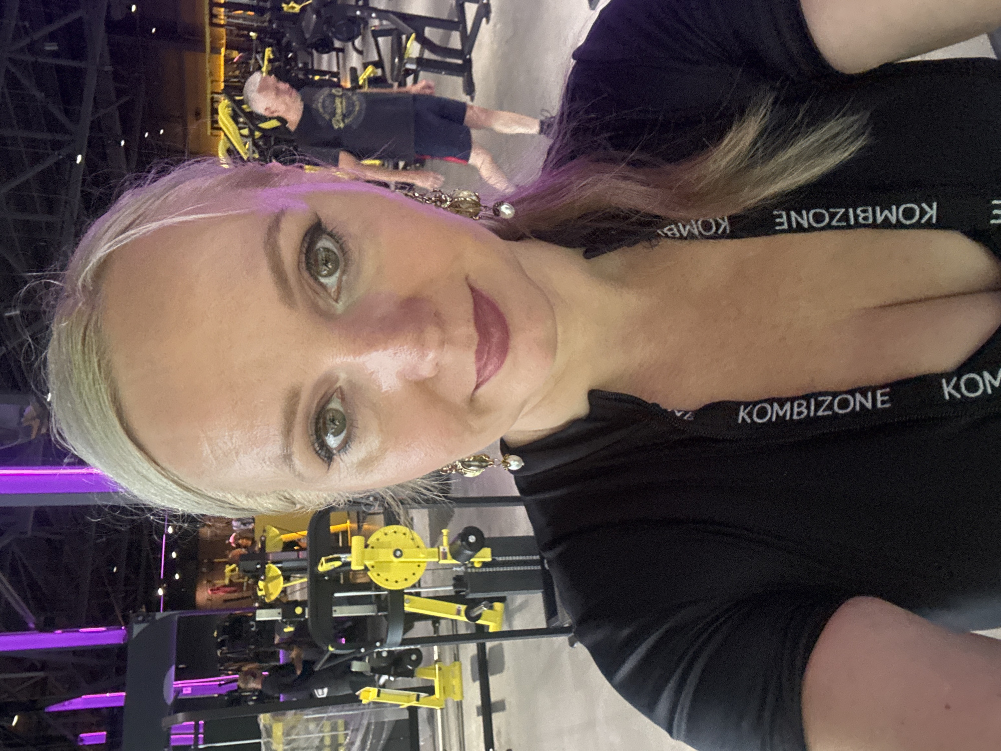 In the city of Komsomolsk-on-Amur, Russia, a woman with blonde hair and light-colored eyes takes a selfie in a gym. She is wearing a black top with "KOMBIZONE" printed vertically on a white band. She has on large, dangling earrings. The background shows gym equipment with yellow accents and purple lighting. A man with grey hair is visible in the background.