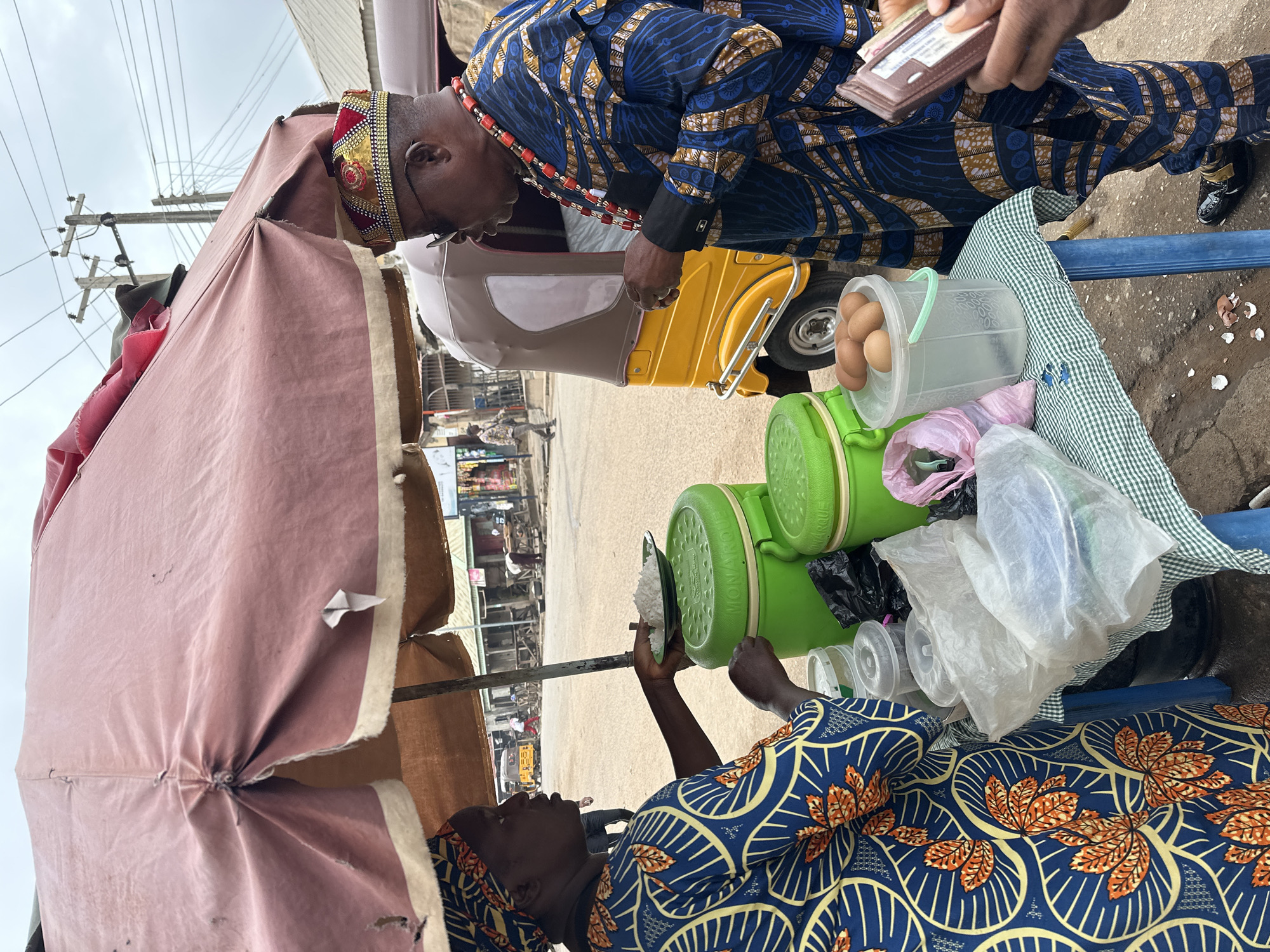 In Narayi, Nigeria, an outdoor market scene unfolds under a pink, tattered umbrella. A woman, adorned in a blue and orange patterned fabric and headwrap, serves a plate of white rice to a man. The man, also in vibrantly patterned clothing of blue and gold, wears a decorative red and gold crown and a beaded necklace. He is looking down at the plate of rice. Beside them, a blue table displays two large green containers, one open and filled with what appears to be rice, and another closed. A plastic bucket on the table holds several brown eggs, and a pile of broken eggshells is scattered on the ground below. In the background, a yellow motorized tricycle, common in Nigerian cities, is parked, and other market stalls and people are visible, suggesting a bustling commercial environment. The sky above is overcast.