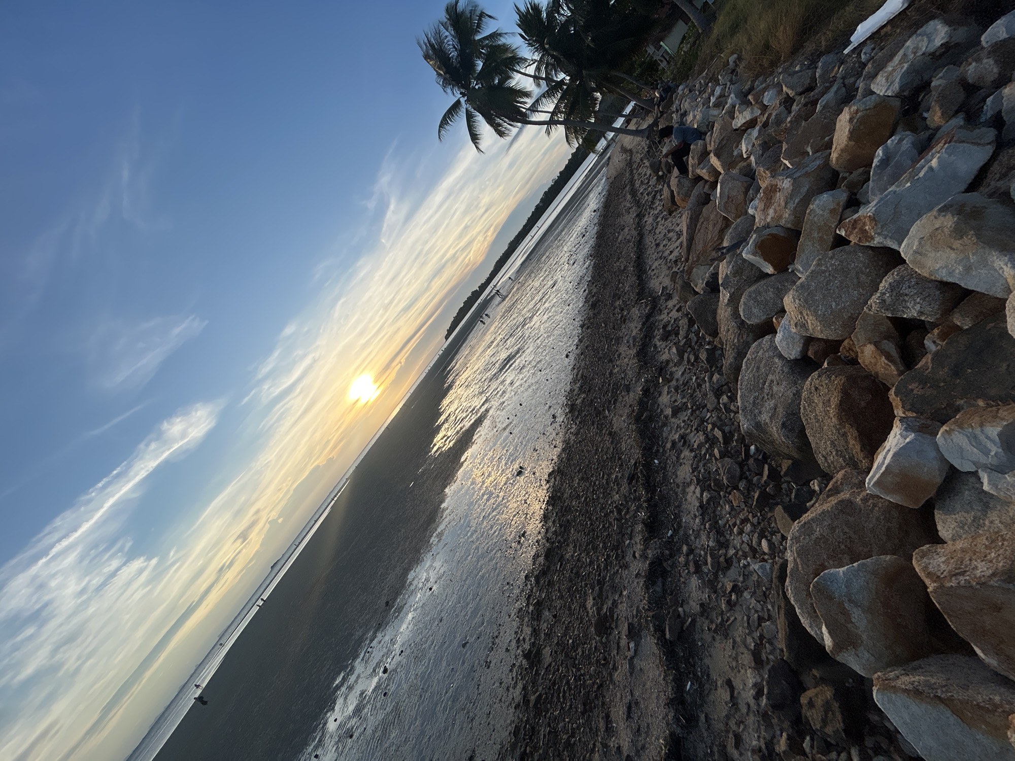 The image shows a beach at sunset in ตำบลเกาะพะงัน, Thailand. The sky is blue with wispy clouds, and the sun is setting on the horizon. The beach is wet, reflecting the sunlight. Rocks and vegetation border the beach. There are some dark palm trees along the beach. There are a few people visible in the distance.