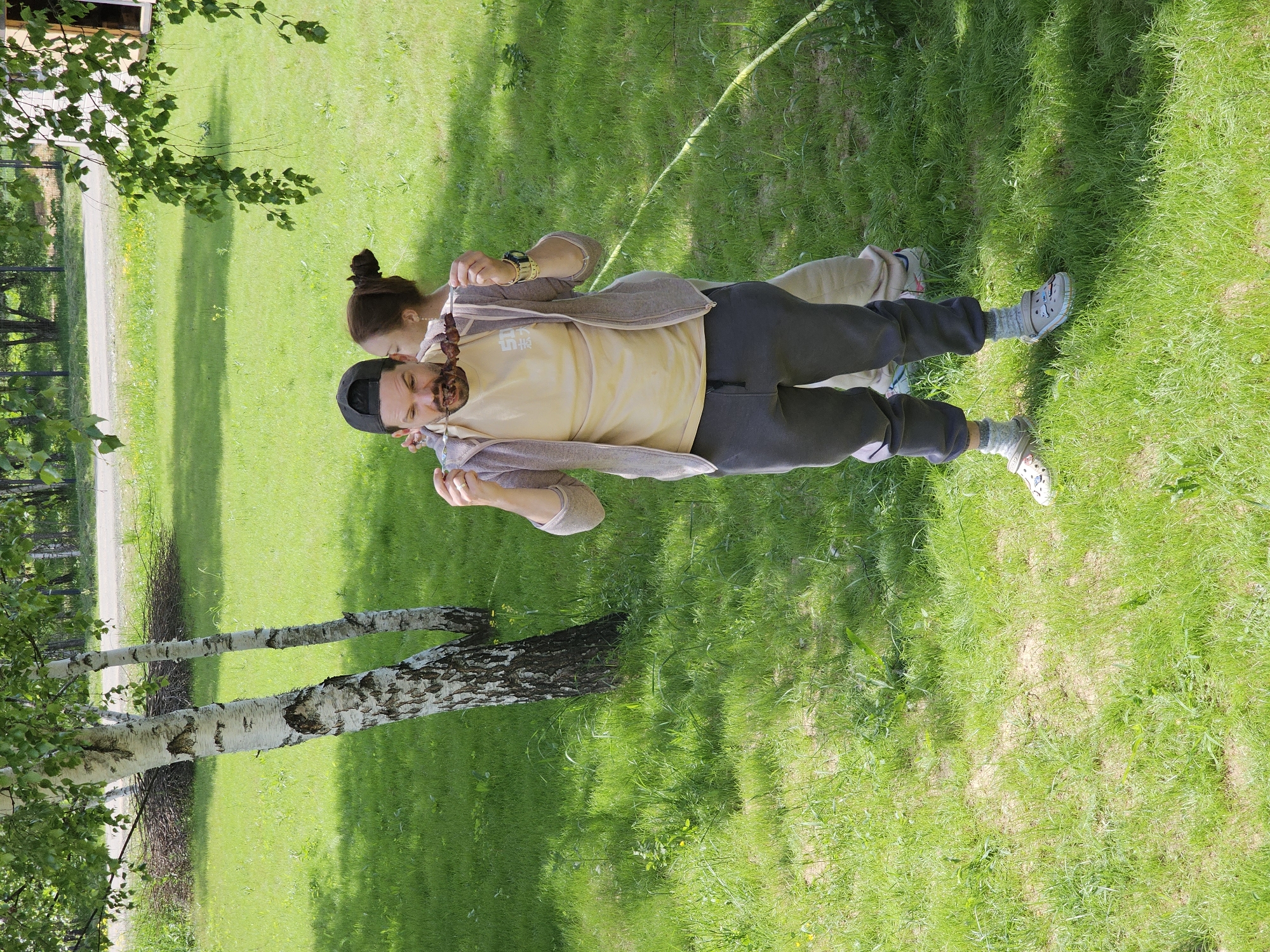 The image depicts a man and a woman standing on a grassy lawn near a birch tree. The man is wearing a black baseball cap, a light yellow T-shirt, a light brown jacket, and dark grey sweatpants. He is holding a skewer of meat up to his mouth as if he's about to eat it. The woman is standing behind him with her hair in a bun, wearing a similar light brown jacket. They are both on the grass, and their feet are visible, wearing socks and Crocs-style shoes.
The background includes lush green grass, a dirt path, and other trees. The location is likely in Мышкино, Russia, as specified, given that the image was provided under the premise of being in that city. The scene suggests a casual outdoor setting, possibly a park or recreational area, where they are enjoying a snack.