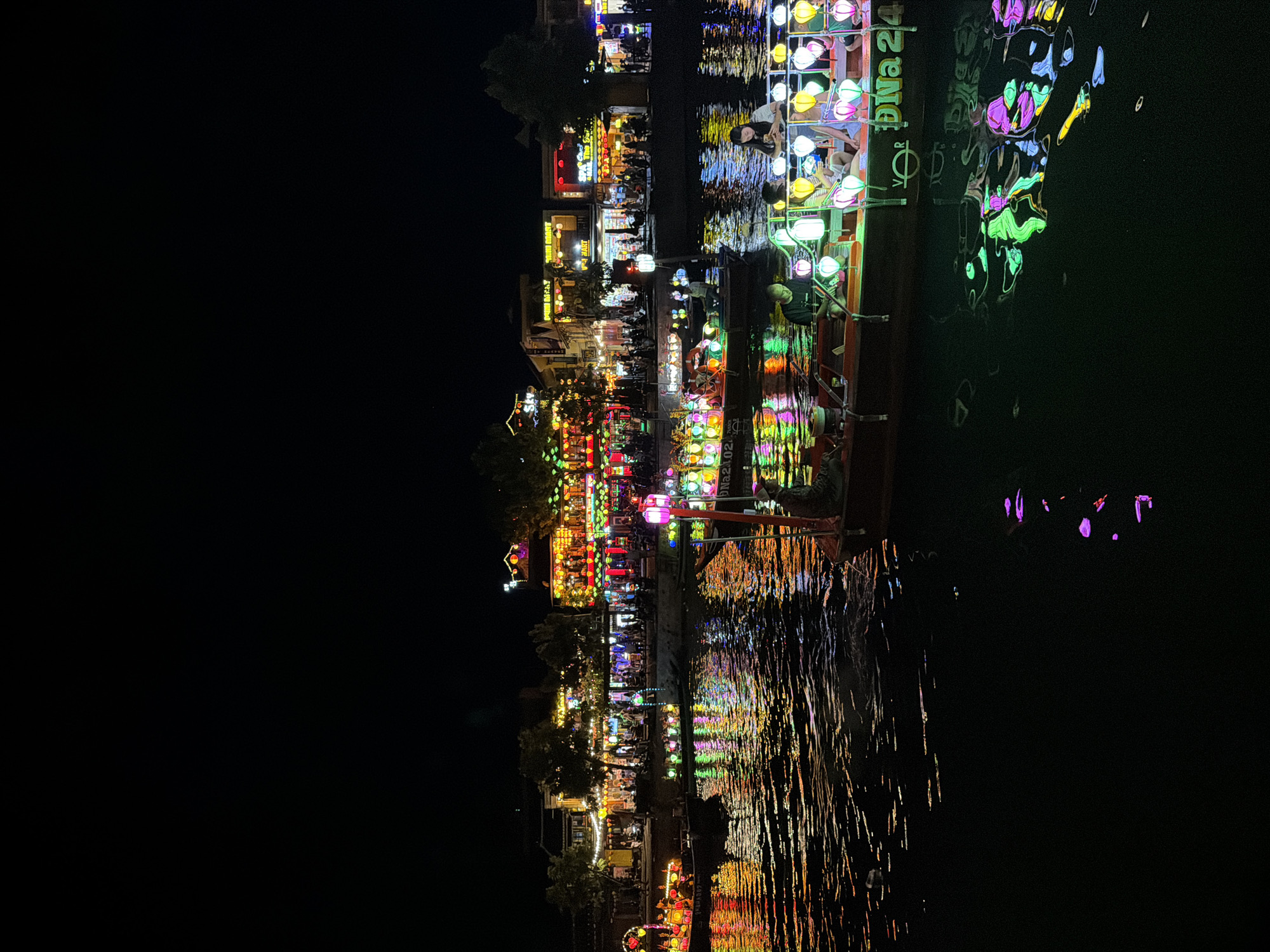 This is a nighttime photograph of the Thu Bon River in Hoi An, Vietnam. The riverfront is lined with multi-story buildings adorned with numerous brightly colored lanterns, casting a vibrant glow on the water. Several boats are navigating the river. One boat in the foreground is decorated with a profusion of colorful lanterns, some of which are lit. There are people on this boat, including a person seated and looking at a phone, and others who appear to be passengers. Another boat, further back, is also illuminated with lanterns. The water reflects the colorful lights from the buildings and boats, creating a dazzling display. The sky is dark, indicating it is nighttime. The scene captures the lively atmosphere of the river at night in Hoi An.