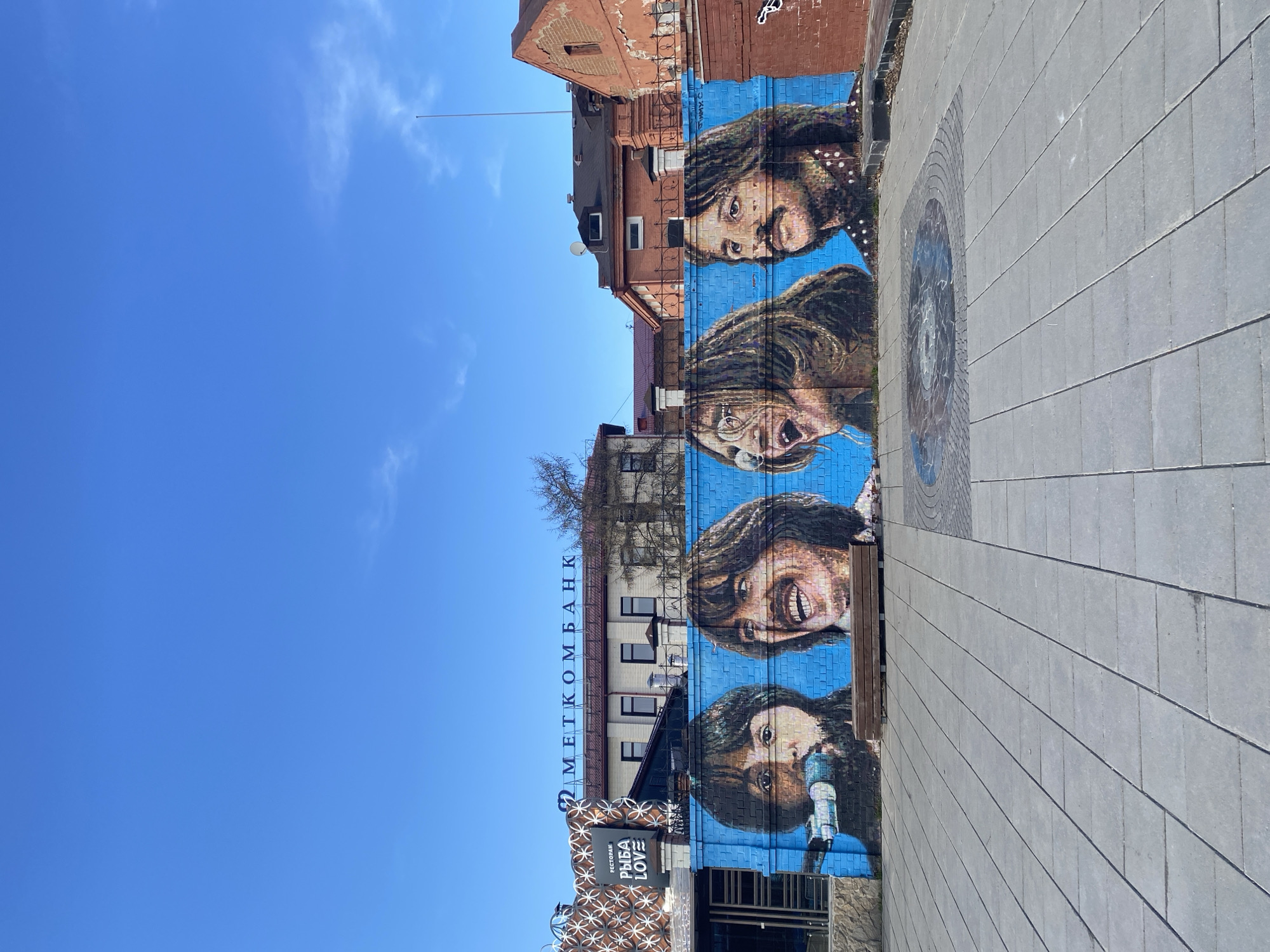 The image shows a building in Yekaterinburg, Russia, featuring a mural of The Beatles on a blue brick wall. The mural depicts the four members of the band, stacked vertically, with their faces prominently displayed. The building also features the text "МЕТКОМБАНК" in Cyrillic script. Below the mural is a restaurant sign that reads "РЫБА LOVE." The ground is paved with gray rectangular stones, with a circular design in the pavement. The sky is bright blue and almost clear of any clouds.