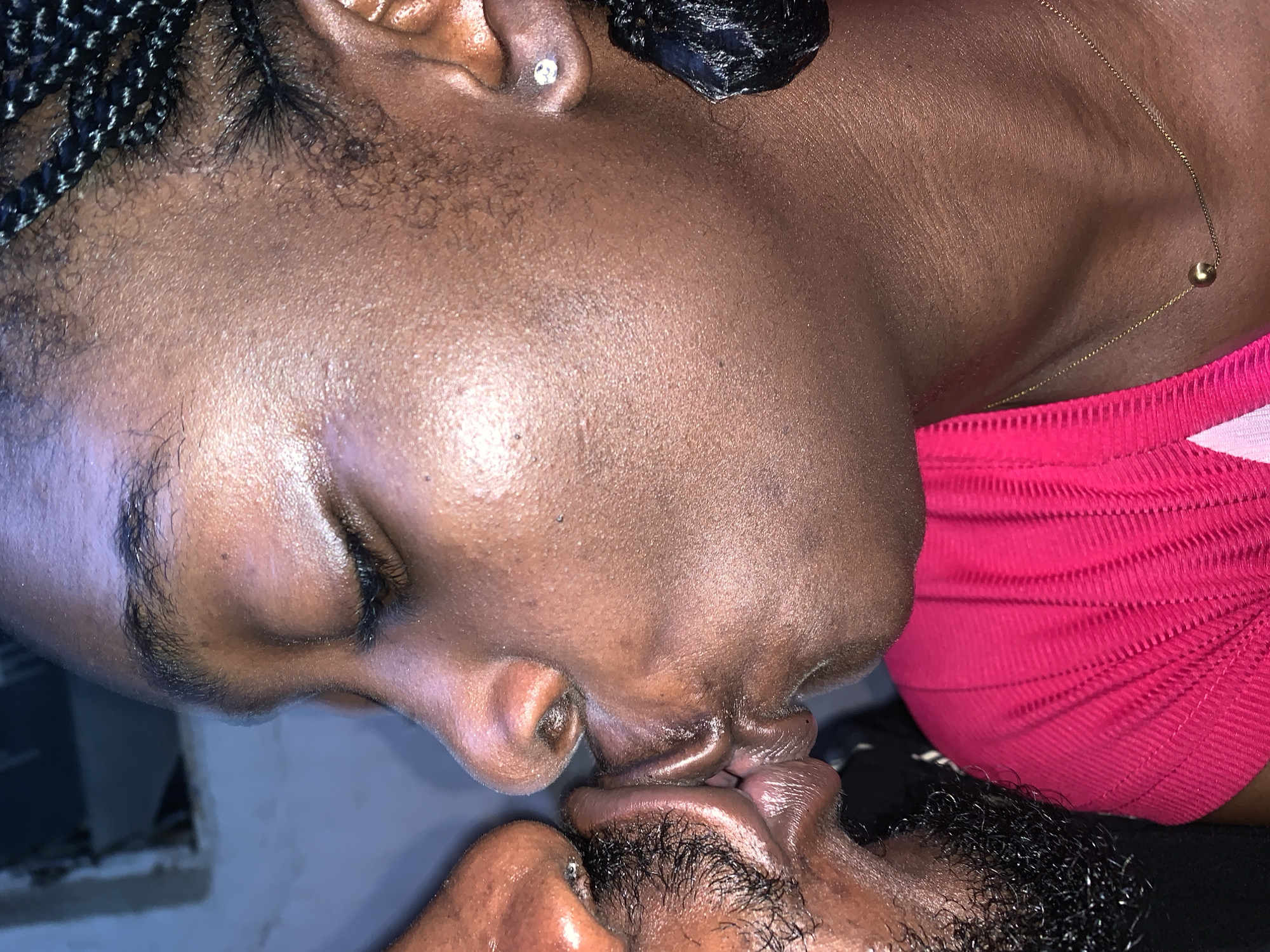 Two individuals are captured in a close-up, profile view, sharing a kiss. The person on the right has their eyes closed and is wearing a bright pink, ribbed top and a delicate gold chain necklace with a single bead. Their hair is styled in dark braids. The person on the left has facial hair and is wearing dark clothing. The background is out of focus, showing a dimly lit interior space with hints of blue and grey tones, suggesting an indoor setting in the city of Rafin Par, Nigeria.