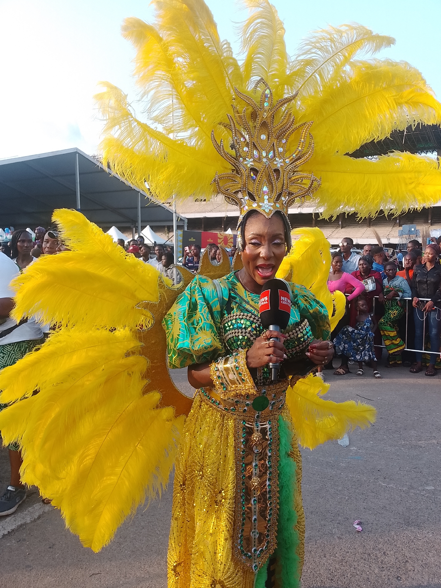 A woman is dressed in a colorful carnival costume with yellow feathers, a jeweled headdress, and a shimmering gold and green dress. She holds a microphone with "News Central" printed on it and appears to be speaking enthusiastically. The background shows a crowd of people, some behind a metal barrier, suggesting she's reporting at an event. The sky is overcast, and there's a large tent structure visible in the background. Based on the carnival attire, the presence of "News Central" and the visual context, this likely depicts the Lagos Carnival in Lagos, Nigeria.