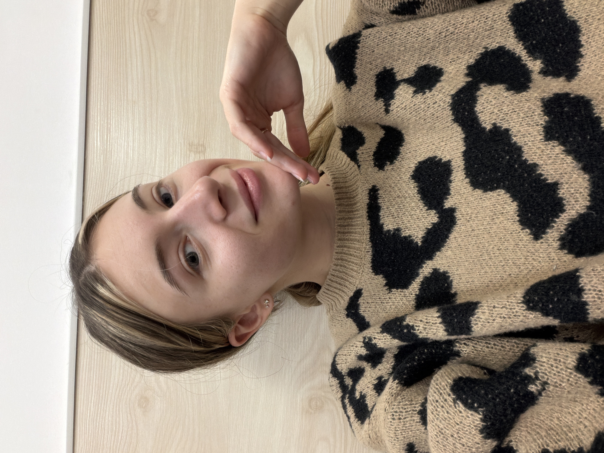 A young woman with light skin and blonde hair, partially highlighted, poses for a selfie indoors. She is wearing a tan and black leopard-print sweater. Her right hand is raised and touching her face near her cheek. She has a small earring in her left ear. The background consists of a light-colored wooden-patterned wall and a white wall in the upper left corner. There is no information to determine this image was taken in Plovdiv, Bulgaria.