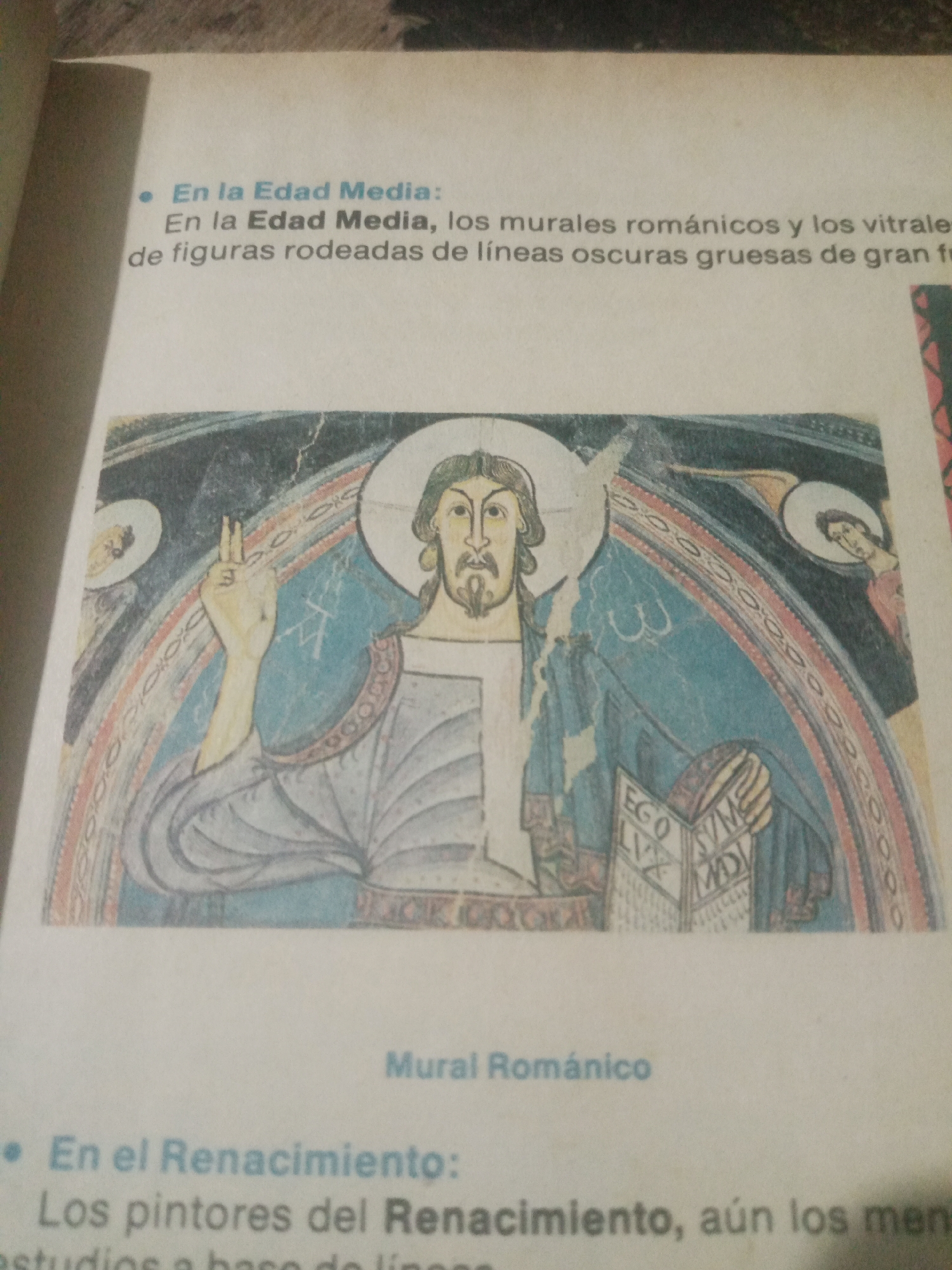 The image displays a page from a book with text and a reproduction of a Romanesque mural. The text discusses art during the Middle Ages and the Renaissance. The mural depicts a central figure, likely Jesus Christ, with a halo, raising a hand in blessing and holding an open book. The figure is dressed in blue and white robes. Two smaller figures with halos are present on either side, possibly angels or saints. The mural features a curved, segmented arch decorated with geometric patterns. Below the mural, the text "Mural Románico" (Romanesque Mural) is visible. The surrounding text, in Spanish, discusses medieval art, murals, and stained glass, and then transitions to Renaissance painters. There is no information in the image that provides context for Santa Ana, Venezuela.