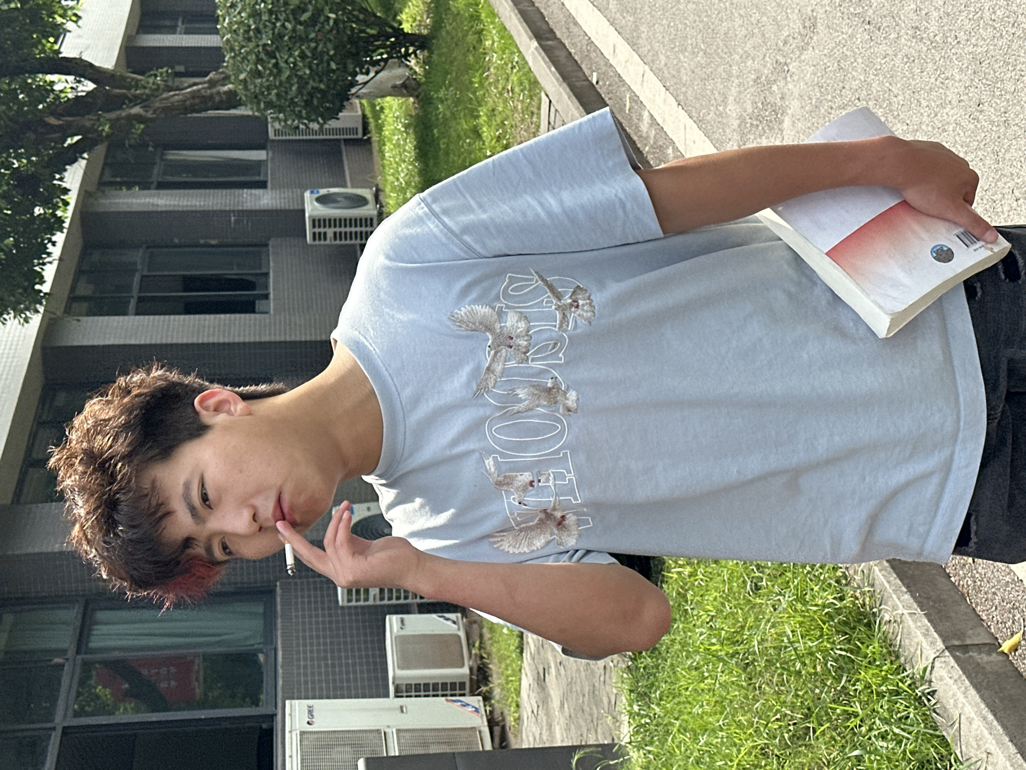 A young man stands outdoors holding a book in his left hand and a cigarette in his right. He has dark, curly hair with a red streak, and he is wearing a light blue t-shirt. Behind him is a multi-story building with visible air conditioning units on the exterior. The surrounding environment includes greenery, grass, and a paved area. The location may be in the city of Jiangjin, China, based on implied information.