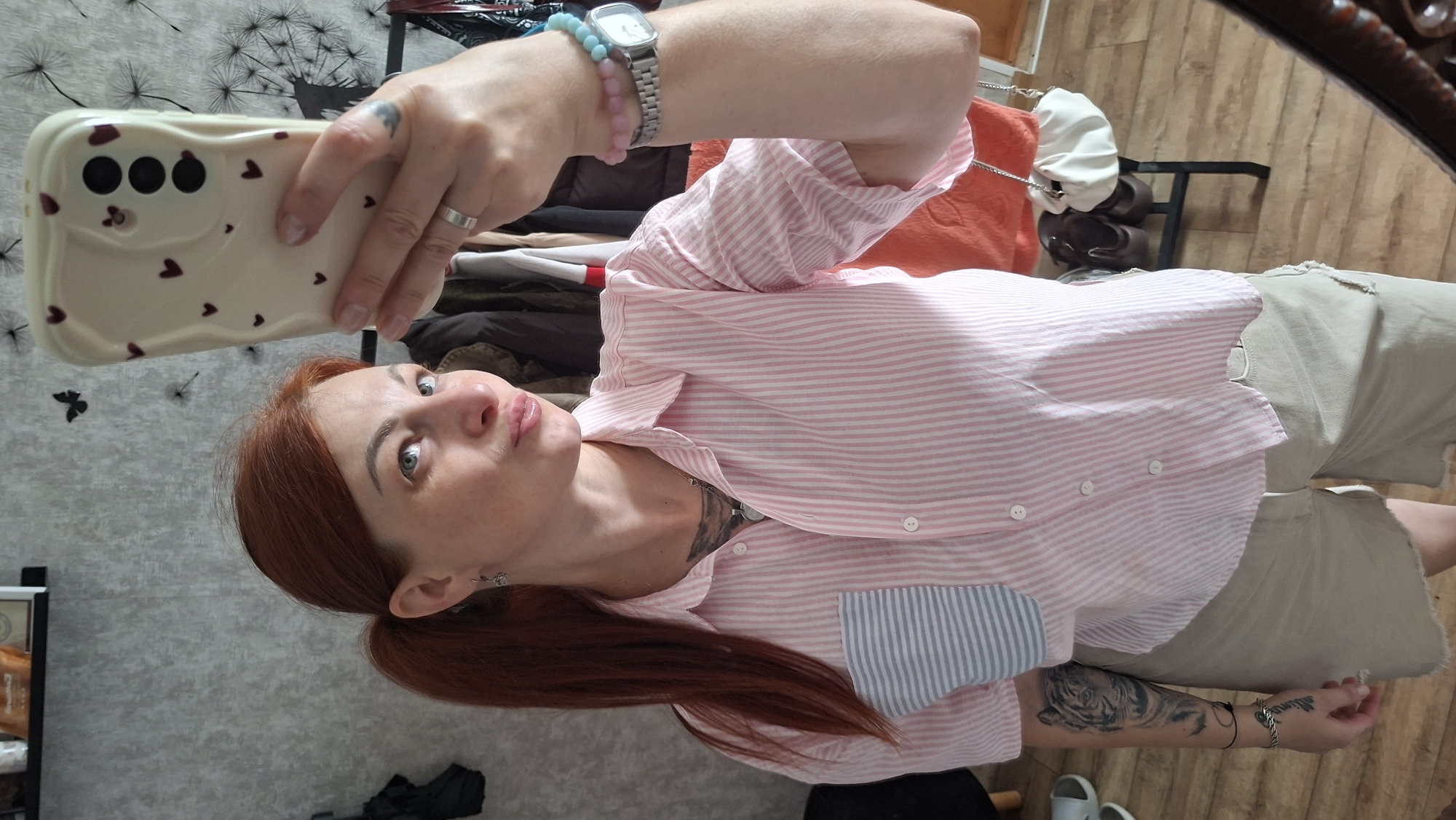 A red-haired woman with a ponytail is taking a mirror selfie in what appears to be a bedroom or dressing area. She is wearing a pink and white striped button-down shirt with a blue and white striped pocket and khaki shorts. She has tattoos on her left arm and neck. On her left wrist, she wears a silver bracelet, and on her right wrist, she wears a silver watch and two beaded bracelets, one pink and one blue. She is holding a white phone case with hearts on it. In the background, there is a closet with clothes, a shelf with items, and a shoe rack with shoes and bags. The environment suggests an indoor setting within a residential space. There is no visual information in the image that indicates the specific location in Myadzyel, Belarus.