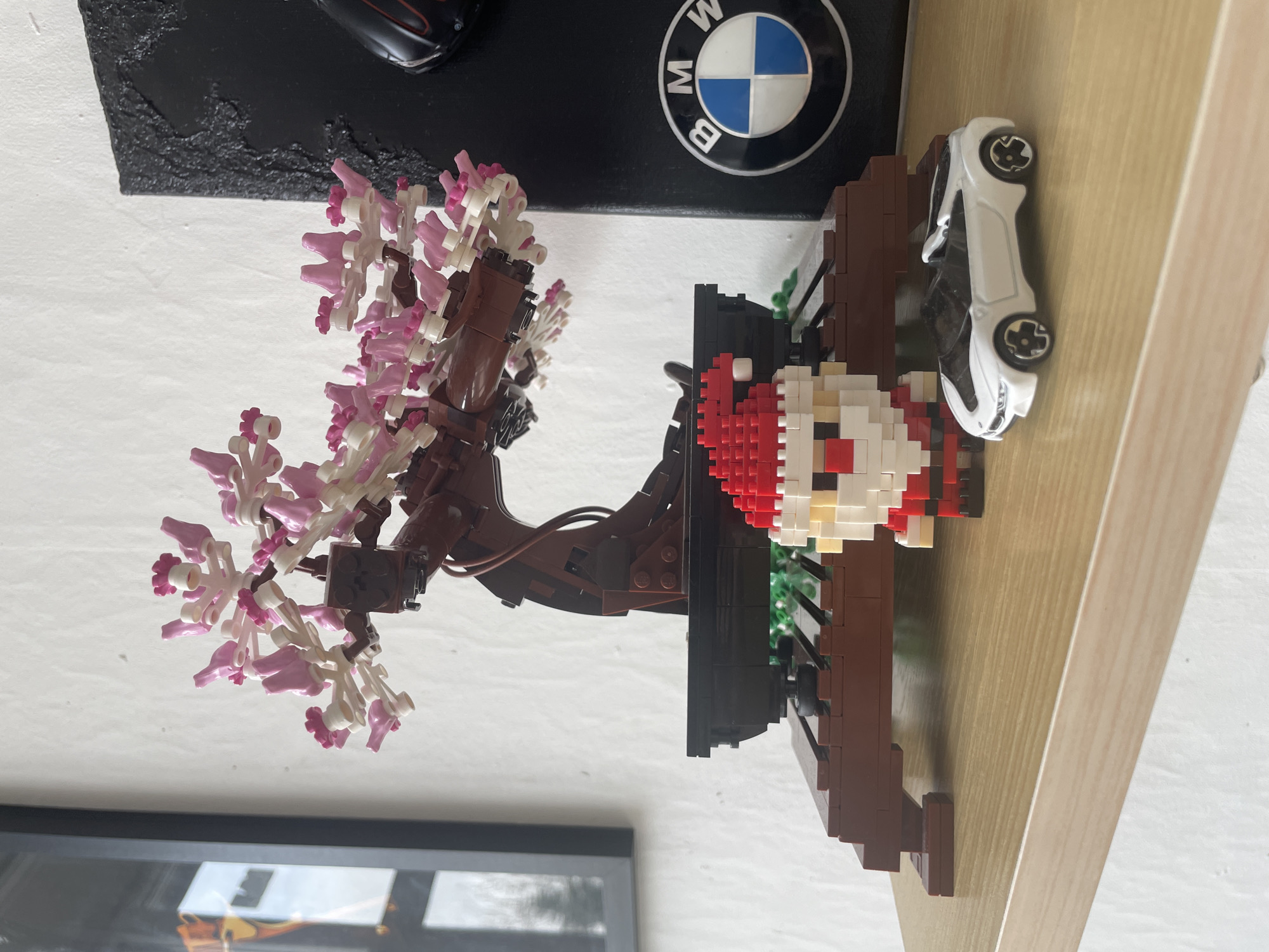 The image shows a shelf with various items. On the left, there is a Lego bonsai tree with pink and white flowers. Above the tree, there is a dark surface with what appears to be a black car model and a BMW logo. To the right of the tree, there is a Lego build that appears to be a character or a scene. Next to it is a white toy car. The shelf itself appears to be made of light-colored wood. The background includes a framed picture.
I am unable to include location information for Sofia, Bulgaria because this information is not in the image.