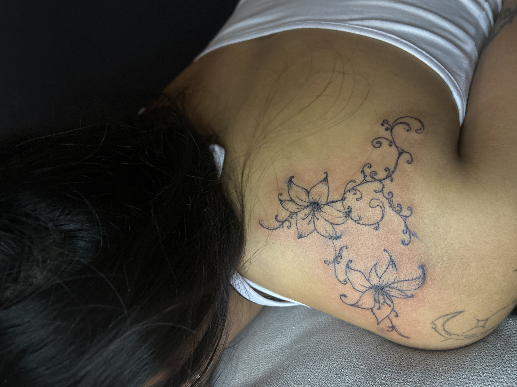 A close-up shot focuses on a person's upper back and shoulder, revealing two flower tattoos connected by a vine design. The person lies on a gray textured surface, and part of their dark hair is visible on the left side. They are wearing a white tank top. A crescent moon tattoo is also visible on the shoulder.
The location is unknown.