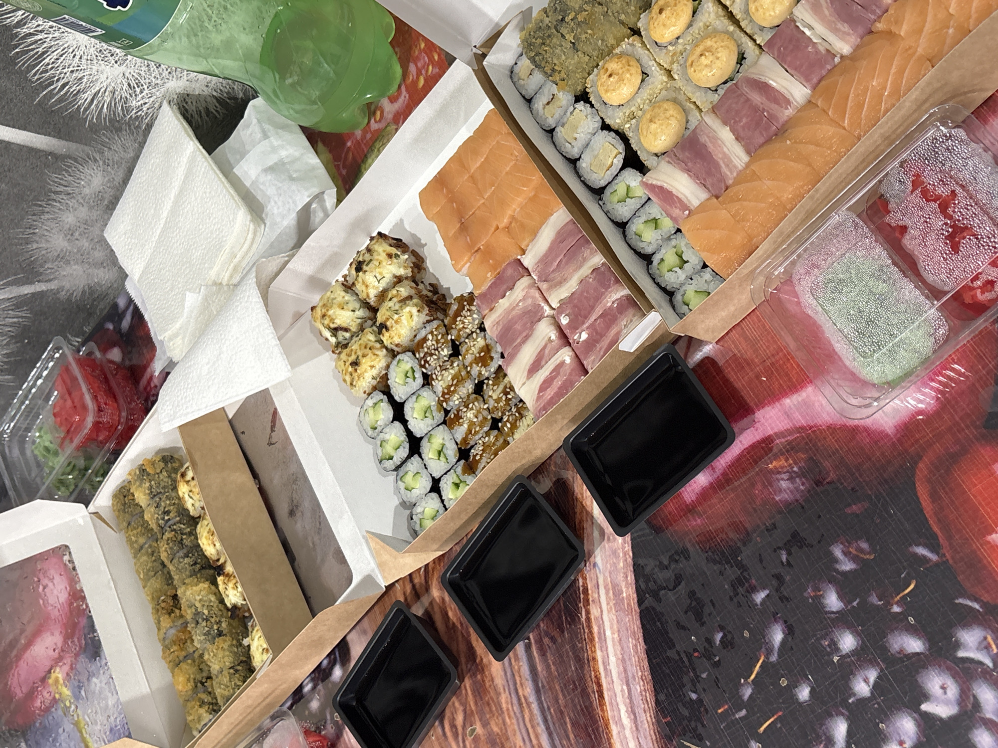 The image shows a spread of sushi in various takeout containers. There are different types of sushi rolls, including some with salmon, bacon, and other toppings. There are also containers with wasabi and ginger. Several small, black square containers, likely for soy sauce, are present. The setting appears to be indoors, possibly on a table with a patterned surface that mimics fruit. There is no specific indication that the image was taken in Rusalevka, Russia, nor is there any indication of human presence.
