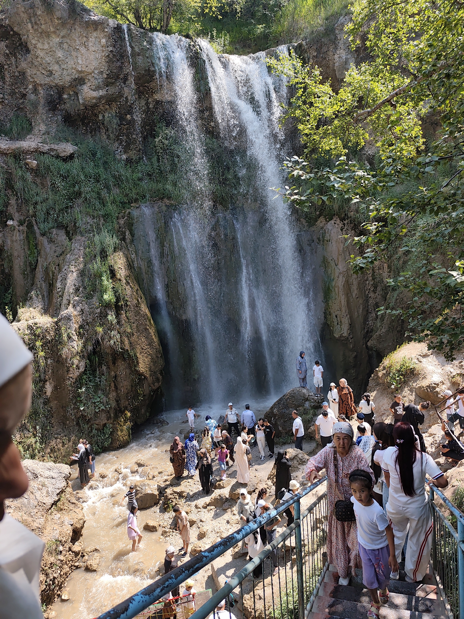 In Arstanbap, Kyrgyzstan, a picturesque waterfall cascades down a rocky cliff face, surrounded by lush green vegetation and trees. A crowd of people, dressed in a mix of traditional and modern attire, gathers at the base of the waterfall and along a set of stairs leading down to it. Some individuals are wading in the shallow water, while others stand on rocks, observing the natural spectacle. The scene is bathed in sunlight, creating a vibrant and lively atmosphere. The waterfall appears to be a popular tourist destination, as evidenced by the number of visitors present.