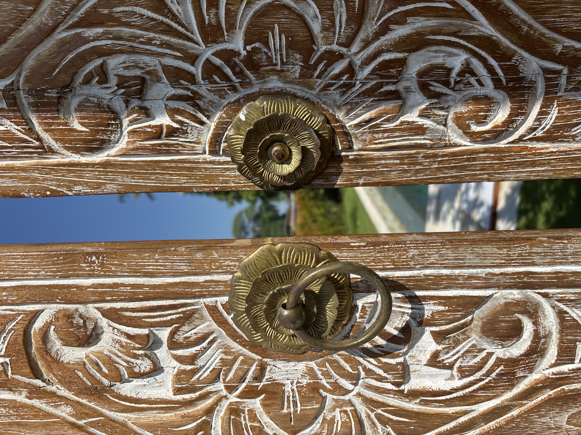 The image shows a close-up of a decorative wooden chest or cabinet door in Cemagi, Indonesia. The wood is light brown with white-washed carving details. Two bronze flower-shaped handles adorn the door, the lower one with a hanging ring. In the background, visible through the space between the doors, is a blue sky and some greenery, suggesting an outdoor setting.