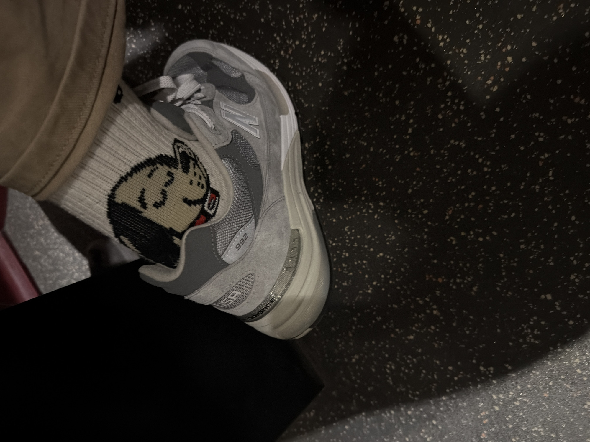The image shows a person's leg and foot, with a focus on their footwear. The person is wearing tan-colored pants and a gray sock featuring a cartoon character. The sock is paired with a gray New Balance 992 shoe. The background is a speckled, dark gray surface. The person is sitting on a black surface. The location is not discernible from the image.