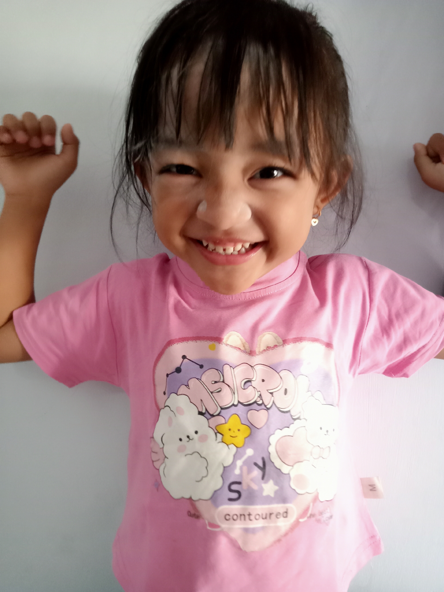 A young girl, possibly of Indonesian descent and likely located in Simo, Indonesia, is shown from the chest up, smiling brightly at the camera. She is wearing a pink t-shirt with a graphic design featuring cartoon characters and text that appears to read "MERCIRO" and "contoured." Her hair is dark and falls forward, framing her face. She has small, heart-shaped earrings. The background is a plain, light-colored wall. Her hands are raised to her sides, fists clenched, in a playful pose. There are white marks on her forehead and nose, possibly from powder or cream.