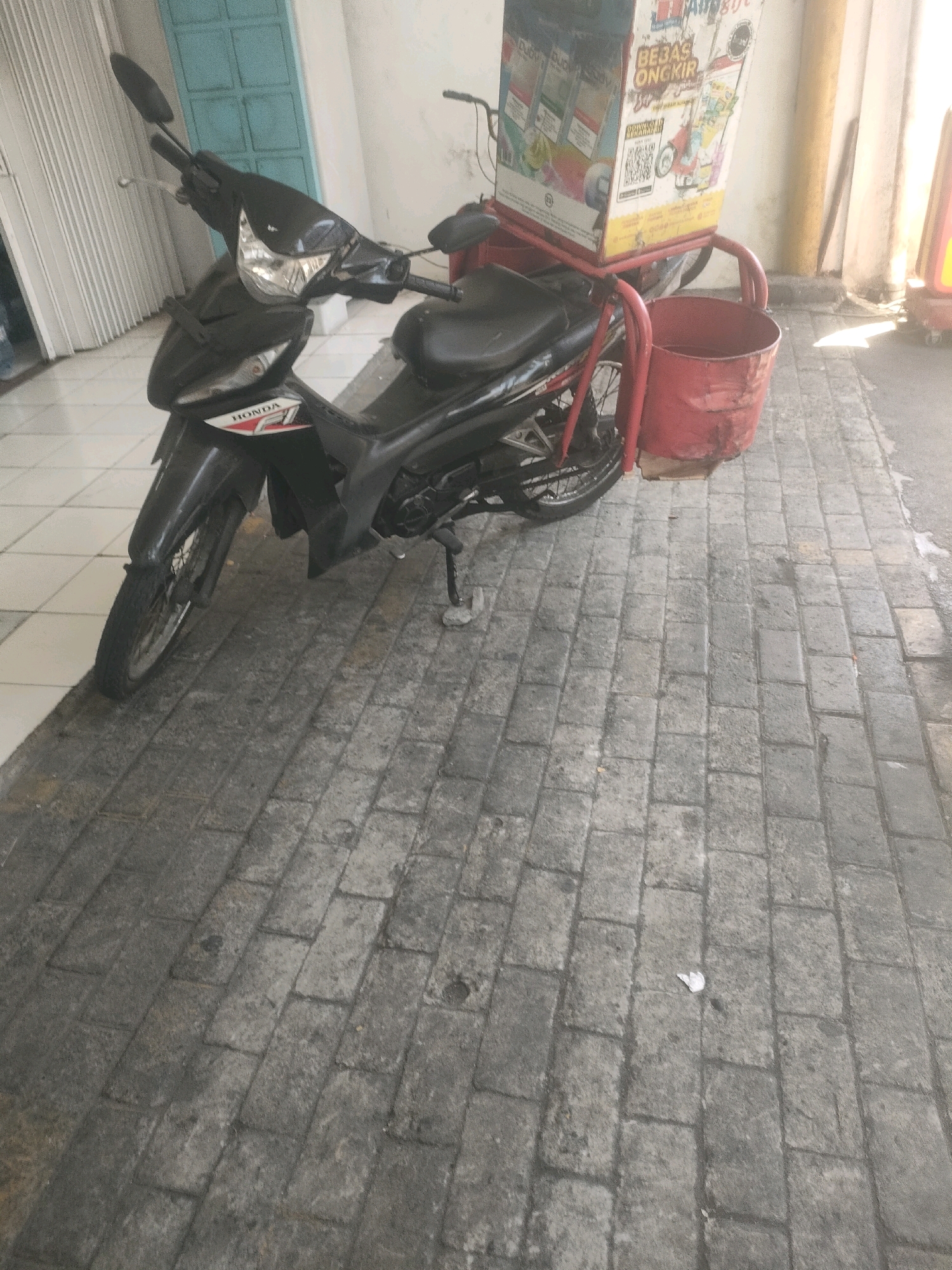 A black Honda motorcycle with a red delivery box is parked on a gray brick sidewalk. The motorcycle has a visible "HONDA F1" emblem. The delivery box is tall and rectangular, with various graphics and text printed on it. Attached to the rear of the motorcycle is a red metal basket. The scene appears to be outdoors, possibly in a commercial or residential area in Surabaya, Indonesia, indicated by the brickwork and building structures visible in the background. The lighting suggests it is daytime.