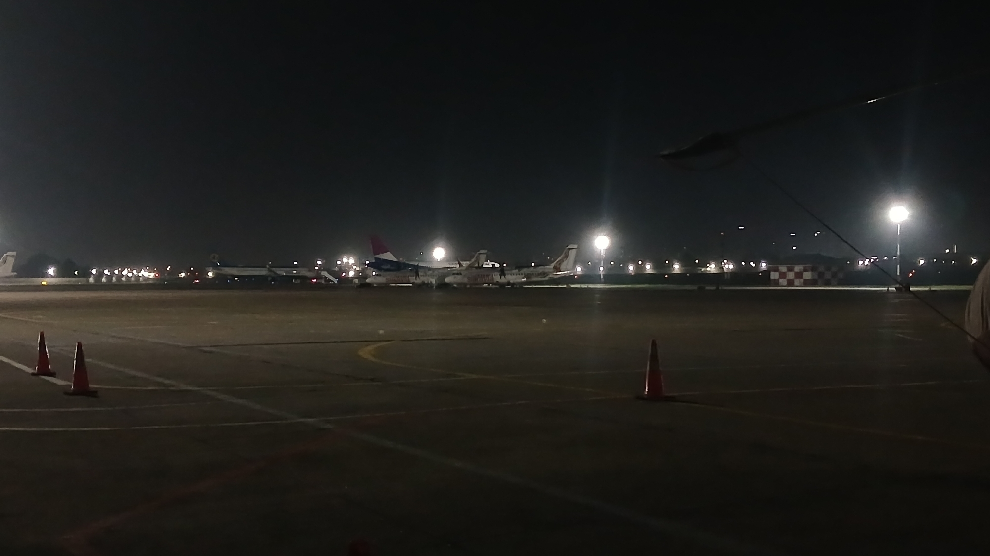 A nighttime airport tarmac scene is depicted with several airplanes parked in the distance, illuminated by bright runway lights and building lights. In the foreground, there are three orange traffic cones on the dark asphalt. The sky is black and starless. The overall impression is one of an operational airport at night. The location is Tashkent, Uzbekistan.