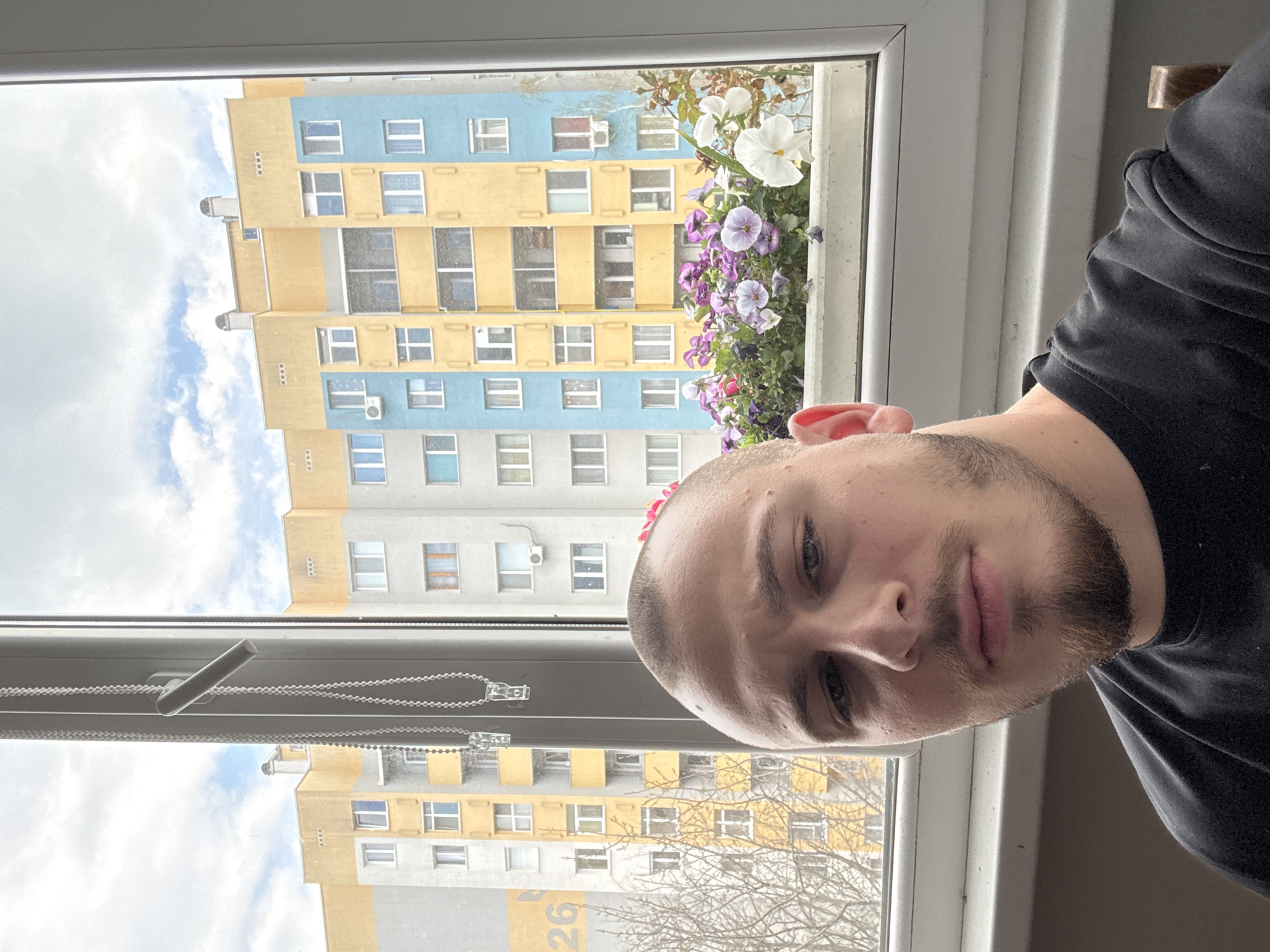 A selfie of a person with short hair and a beard, wearing a black shirt, is taken indoors near a window. The window overlooks a residential building with a mix of yellow, blue, and grey facade colors. Flowers in a window box are visible on the window sill. The photo was taken on a cloudy day. Based on the architectural style of the buildings and other contextual clues, it is likely the location is Stara Zagora, Bulgaria.