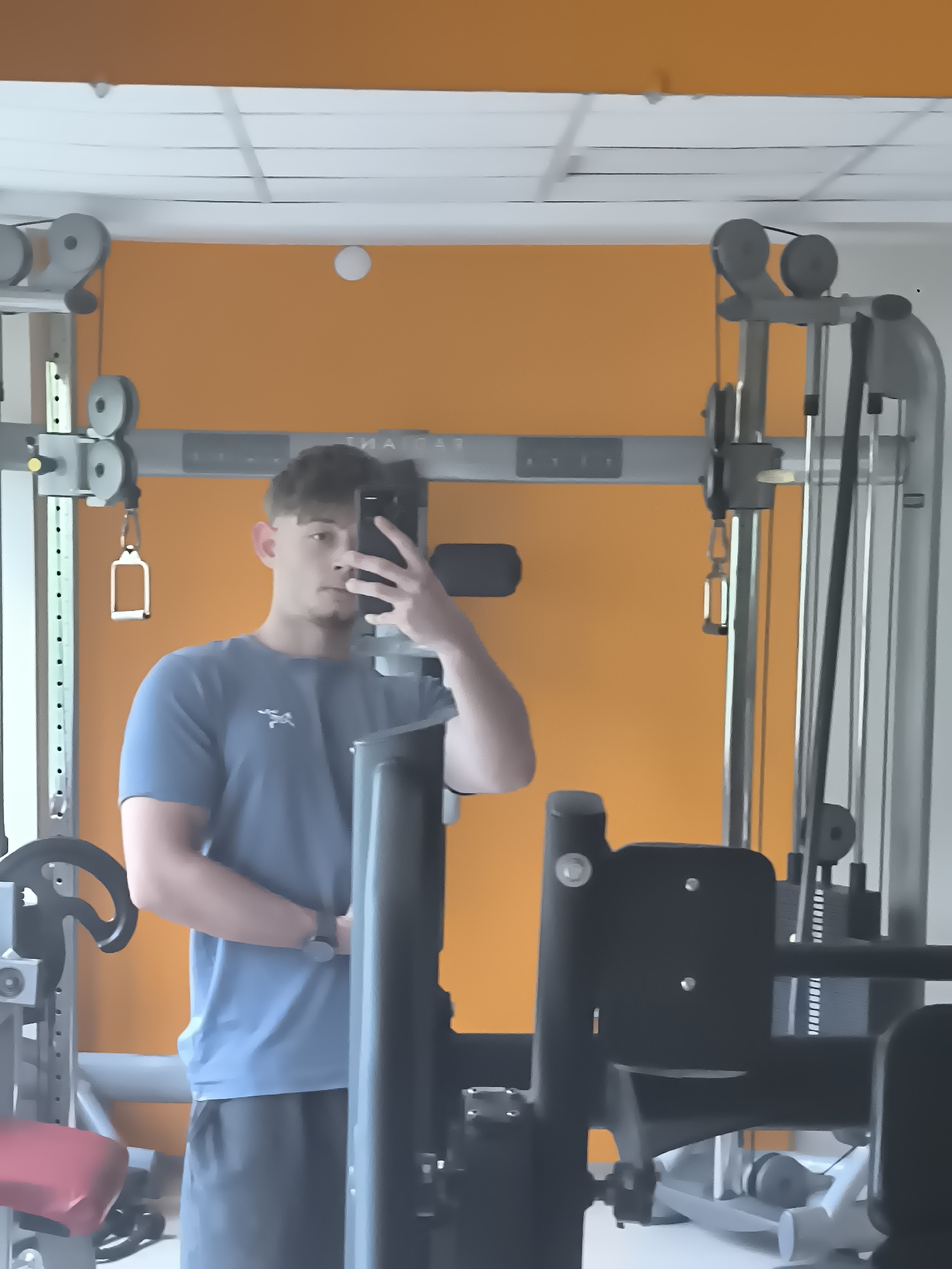 A young man is taking a selfie in a gym. He is wearing a blue t-shirt and black shorts. The gym has orange walls and weight machines. The location is likely in Sofia, Bulgaria.