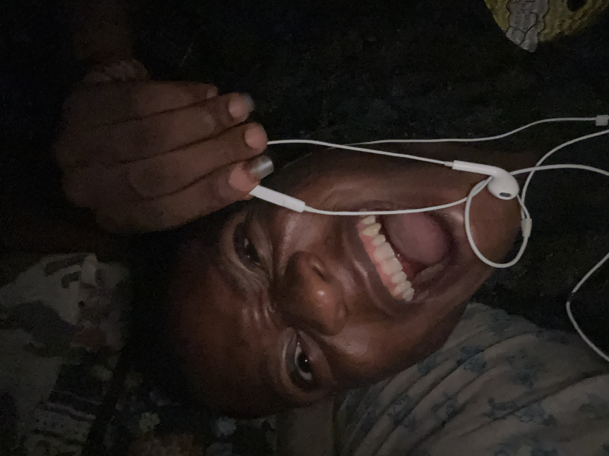 A dark-skinned person is lying on their back with their head tilted to the right. The person is smiling widely, with their mouth open. They have white earbuds in their ears. One of their hands is near their head. They appear to be lying on a bed with floral patterned bedding. The location and city are unknown.