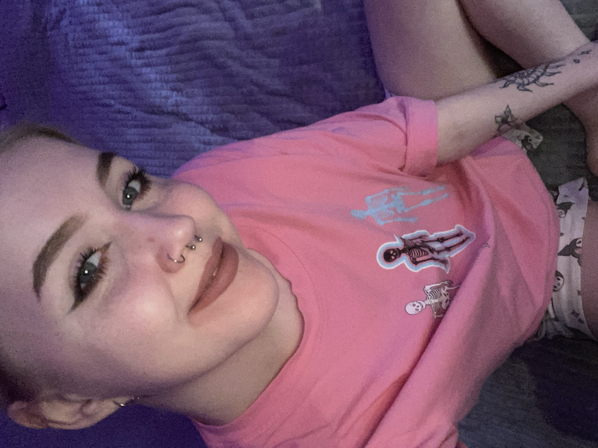A woman is lying down on a blue blanket. She is wearing a pink t-shirt with skeleton graphics. She has fair skin, light brown eyebrows, green eyes, a nose piercing, and small hoop earrings. Her left arm is visible with tattoos on the forearm. Part of her patterned white shorts is visible. The location is unspecified.