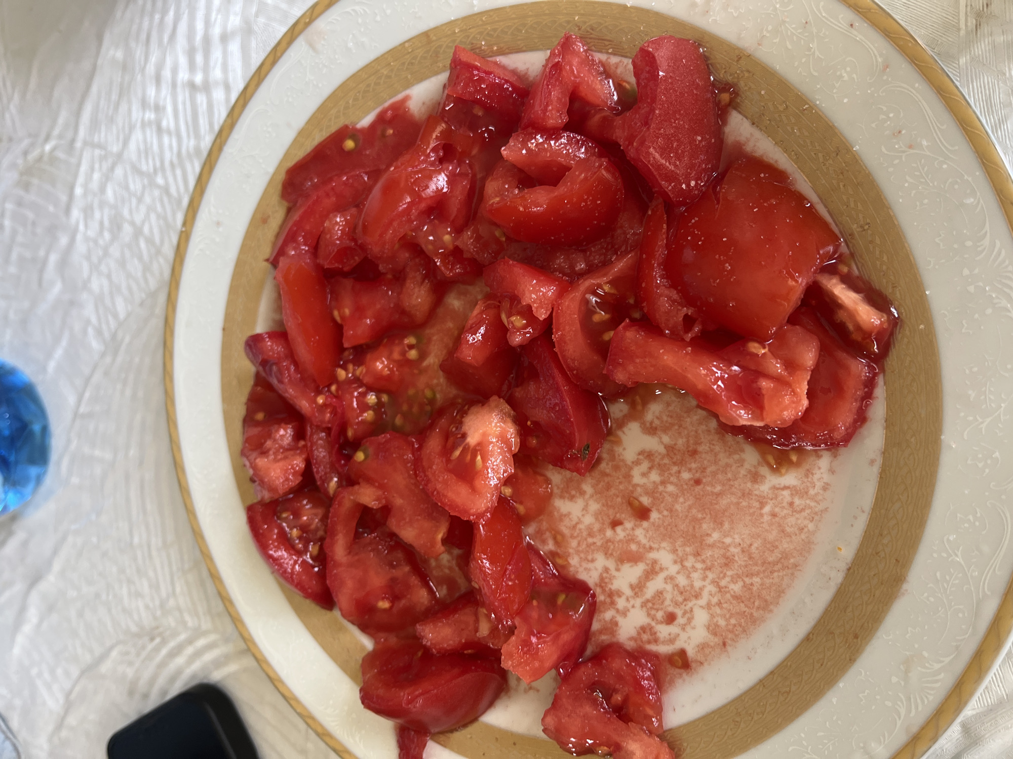 A plate of chopped tomatoes, some seeds visible, sits on a decorative white plate with gold trim. The plate is positioned on a white surface. A blue object is partially visible in the upper left corner, and a black object is in the lower left corner. The scene does not contain enough information to establish a location in Zlatograd, Bulgaria.