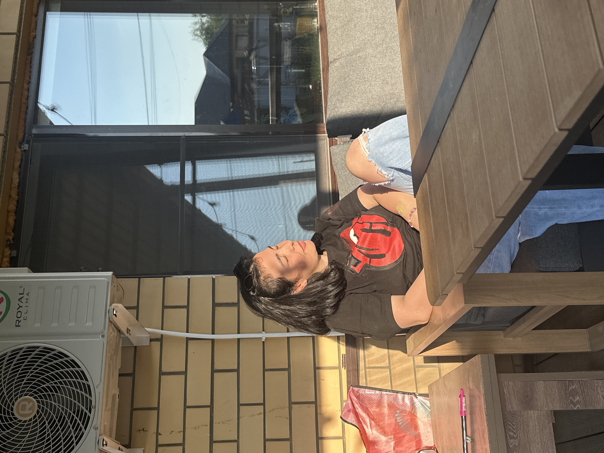 A woman sits at a picnic table in an outdoor setting in Unknown, Unknown. She wears a black shirt with a red graphic, light-wash denim shorts, and dark sunglasses pushed back in her hair. The picnic table, made of light-colored wood, occupies the right portion of the frame. The background consists of a brick wall with an air conditioning unit mounted on it, a window, and outdoor furniture. A white cable runs along the wall. A pink and black pen, and a red and white cloth are near the picnic table.