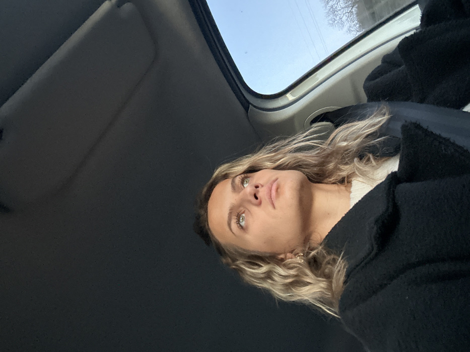 A woman with blonde, wavy hair and green eyes sits in a car. She wears a black coat over a white sweater. The car's interior is visible, including the sun visor and the sunroof, through which a bright, pale sky is seen. Her face is tilted upward, and she gazes off-camera.