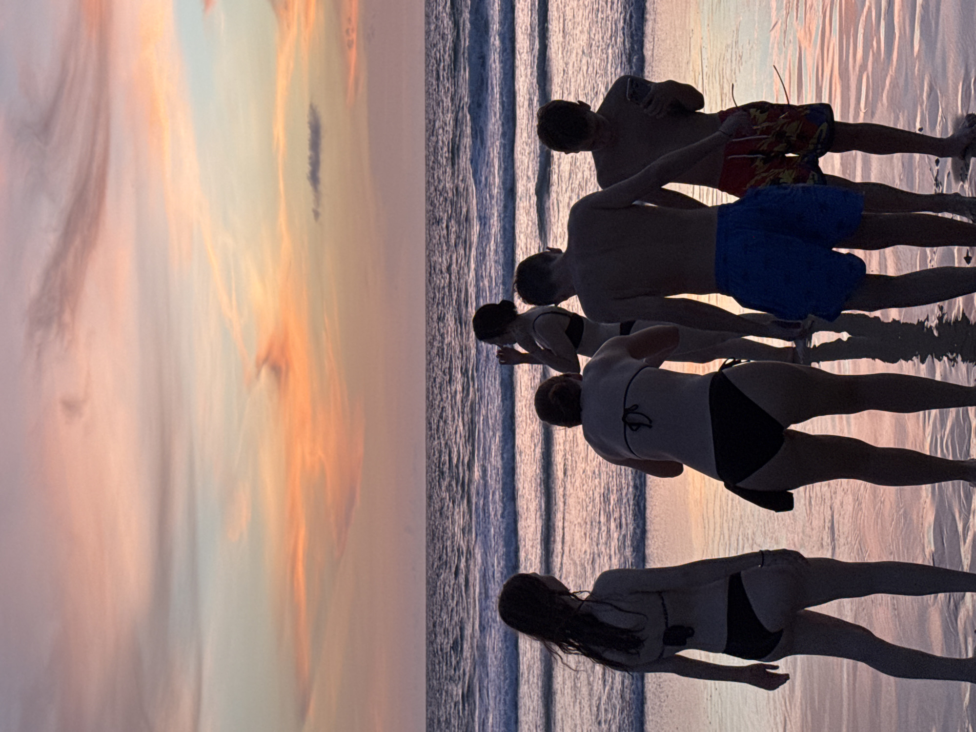 At Karon Beach, Thailand, a group of five young people stand on the wet sand near the ocean's edge as the sun sets. The sky is filled with soft pastel colors of pink, orange, and purple from the sunset. The ocean waves are gently rolling onto the shore, reflecting the sky's colors. The group, silhouetted against the colorful sky, consists of three females and two males. They are dressed in swimwear. One female on the far left is facing away from the camera. Next to her, another female is also facing away, looking towards the ocean. In the center, a third female is captured in profile, her back to the viewer, holding her hand to her face. To her right, a male is facing away, wearing blue swim trunks. To his right, another male is facing towards the viewer's left, holding a smartphone. The sand is wet and reflective, showing the colors of the sunset and the silhouettes of the people.