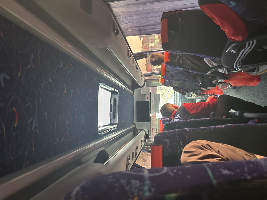 The image depicts the interior of a passenger bus in Aksakovo, Bulgaria, viewed from a rear seat looking forward. The bus features blue and purple patterned seating with red headrest accents. Overhead storage compartments and light fixtures line the ceiling, which is dark blue with abstract colorful patterns. A central roof hatch with a mesh screen is visible. A small monitor is mounted on the front bulkhead of the bus.
Several individuals are seated. In the mid-ground left, a male subject wearing a red and black jacket with the partial word "SPORTIV" visible on the back is seated, looking forward, and holding a red object in his right hand. To the immediate right, another male subject is seated facing forward, seen from behind. On the far right, black athletic pants with white Adidas stripes are visible on a seated individual. Multiple red plastic bags hang from the back of seats along the right aisle. Brown or purple curtains are drawn partially over the windows on the right side.
Through the front windshield, an exterior scene shows a road or track with vegetation on the left side and a partially visible sign with Cyrillic text. Through the right-side windows, residential buildings with tiled roofs and a bright, possibly sunset or sunrise, sky are visible.