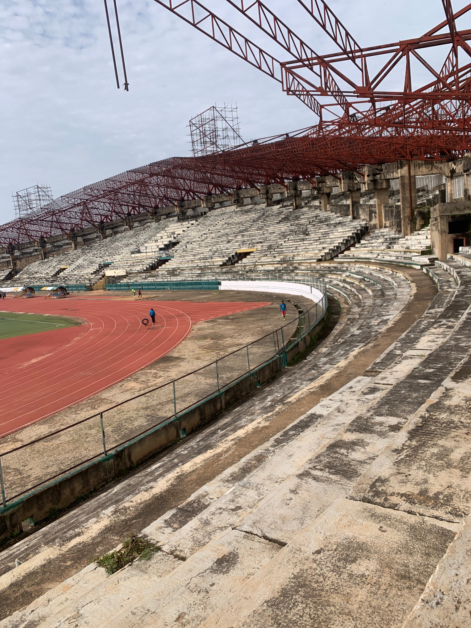 This is a wide-angle, eye-level shot of a section of the newly constructed National Stadium in the Surulere area of Lagos, Nigeria, taken from the upper seating tiers. The stadium is in an advanced stage of construction, with the skeletal red steel framework of the roof structure dominating the upper half of the image. Below the roof, rows of concrete stadium seating stretch outwards and downwards, some sections appearing weathered and stained. A red running track is visible in the lower left quadrant, with green turf beyond its inner edge. Several individuals are present: one person in a blue top is standing on the track near a large tire, another in a red shirt is further away on the track, and two more figures are walking on a path behind the track. Scaffolding is erected in several locations around the stadium's upper levels, suggesting ongoing construction work. The sky is overcast, contributing to a muted light across the scene. The overall impression is one of a large, modern sports facility in progress.