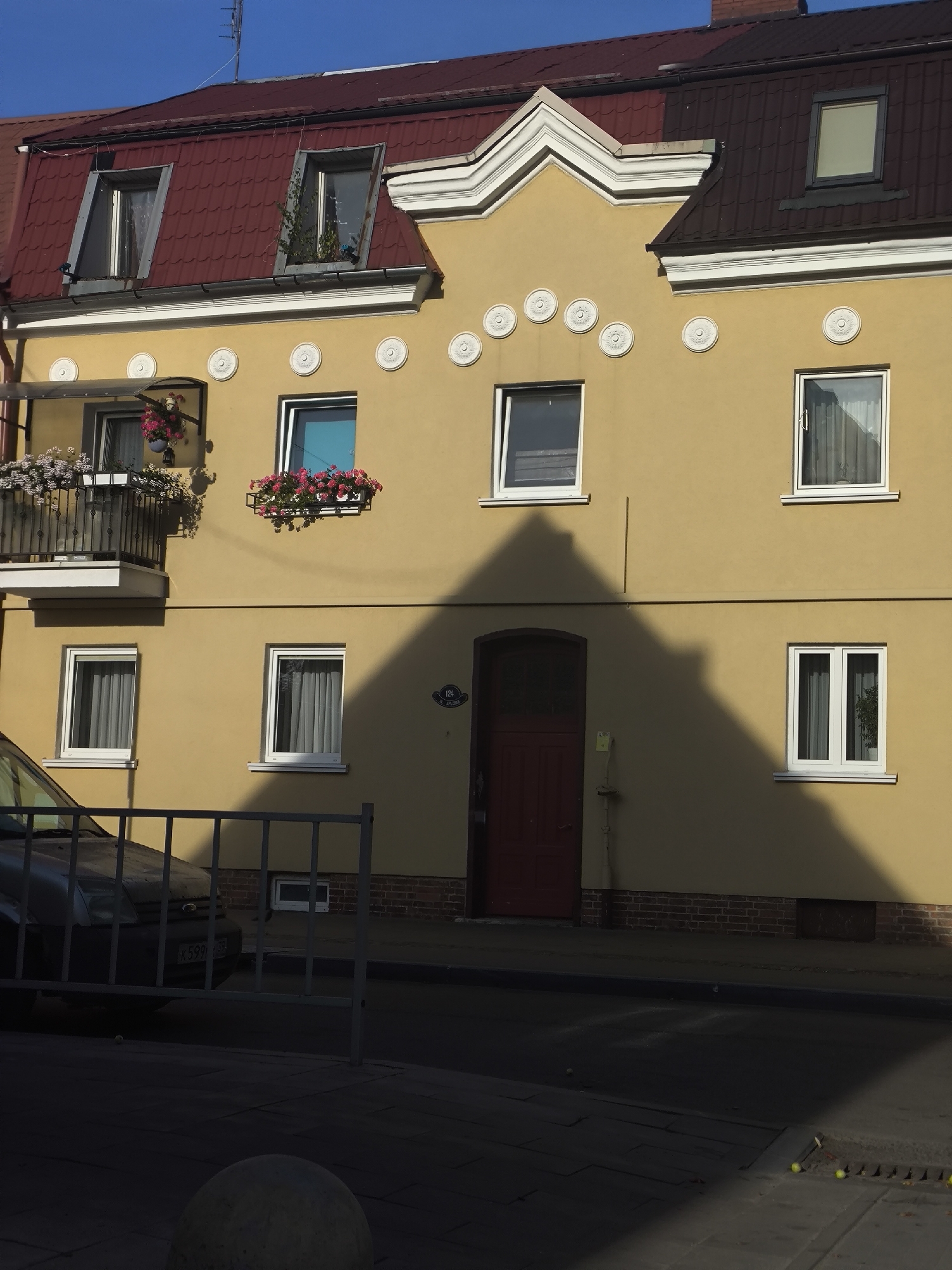 The image depicts the facade of a three-story building in Kaliningrad, Russia, painted in a pale yellow hue. The building features a red-tiled mansard roof with two dormer windows on the upper level. The main facade has several white-framed windows, some adorned with planters containing pink and white flowers. A black metal balcony railing is visible on the second floor. A large, dark red door is centrally located on the ground floor. The building is casting a prominent triangular shadow from a nearby structure onto its facade, indicating a sunny day. In the foreground, a dark-colored car is partially visible behind a grey metal barrier, parked on the street in front of the building. The street itself is paved, with some areas appearing shadowed. The general environment suggests an urban setting.