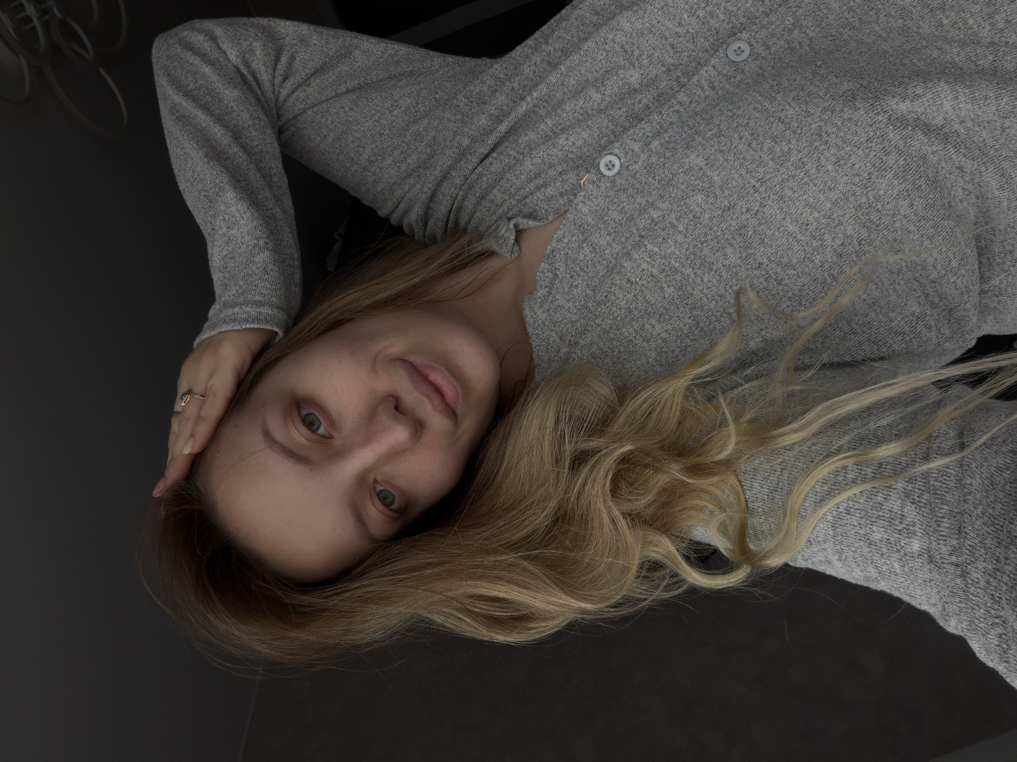 A young woman with blonde hair lies on a dark, possibly black, surface. She's wearing a light grey, button-down cardigan or sweater. Her left hand rests on her forehead. The background is predominantly dark and out of focus. There is no information available that suggests this image was taken in Reshetnikovo, Russia.