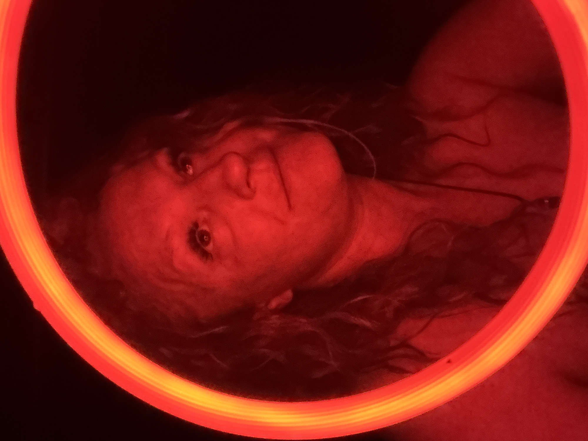This striking portrait, captured in Chaikovsky, Russia, features a person, likely female, framed by a prominent, glowing circular light source. The subject's head and shoulders are visible, positioned within the red and orange light ring. The person has long, wavy, dark hair that partially blends into the very dark, almost black background.
The main activity is a stylized portrait session, with the subject looking directly at the viewer with a neutral to contemplative expression. The strong red and orange artificial light casts dramatic illumination on the subject's face and skin, highlighting features while leaving much of the background in deep shadow. This lighting effect creates an intense and somewhat mysterious atmosphere.
Notable details include the vivid, almost fiery colors of the light ring, which provides the primary illumination for the scene. The skin tone of the subject appears flushed red due to the dominant light. Small wires or a necklace are faintly visible around the subject's neck area, partially obscured by the lighting and shadow. No specific location cues within Chaikovsky, Russia are visible in the image itself, besides the overall low-light and indoor/night setting suggested by the artificial illumination. There is no visible text in the image.