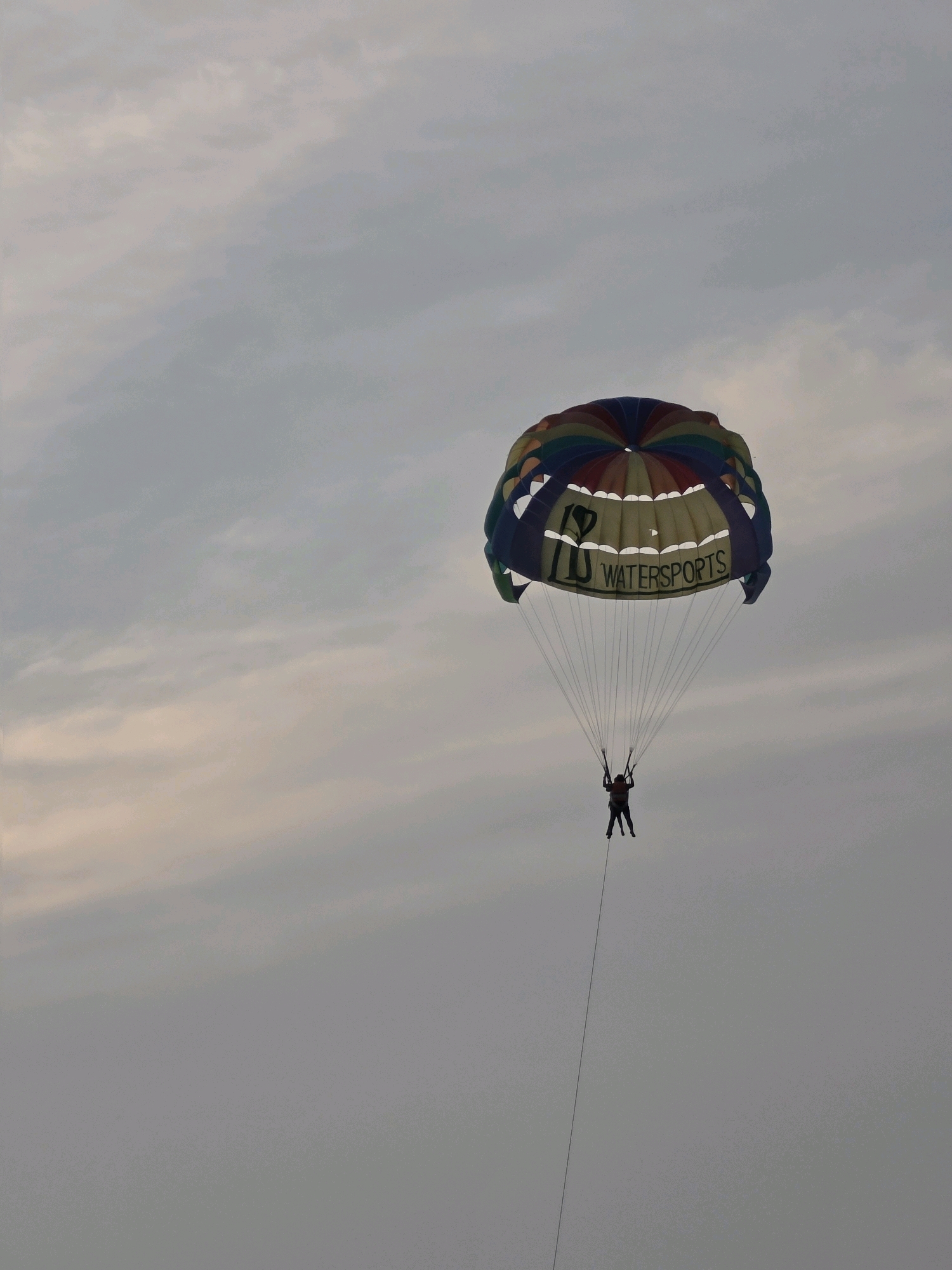 Two individuals are parasailing high in the sky. They are attached to a colorful parachute adorned with the words "WATERSPORTS". The sky is overcast with a mix of grey and pale pink clouds. The scene appears to be near the coast of George Town, Malaysia, given the prevalence of watersports activities in the area.