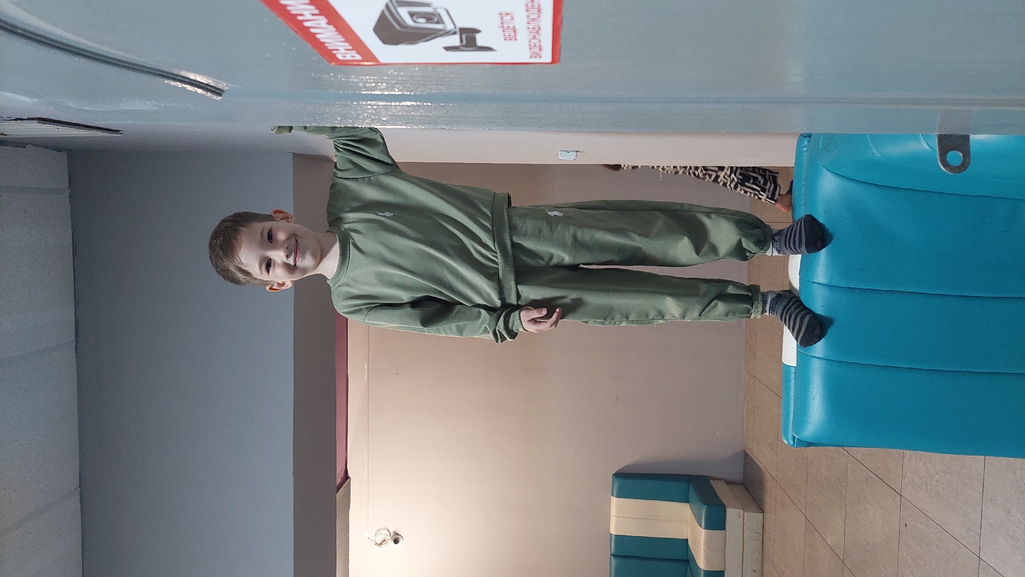 A young boy stands on a bright blue inflatable structure in what appears to be an indoor waiting area. He is wearing a green tracksuit and striped socks. To his right, a door with a red and white "WARNING" sign featuring a security camera is visible. In the background, a light-colored wall, a beige and blue couch, and a tiled floor can be seen. The lighting suggests an artificial indoor environment, possibly in Komsomolsk-on-Amur, Russia.