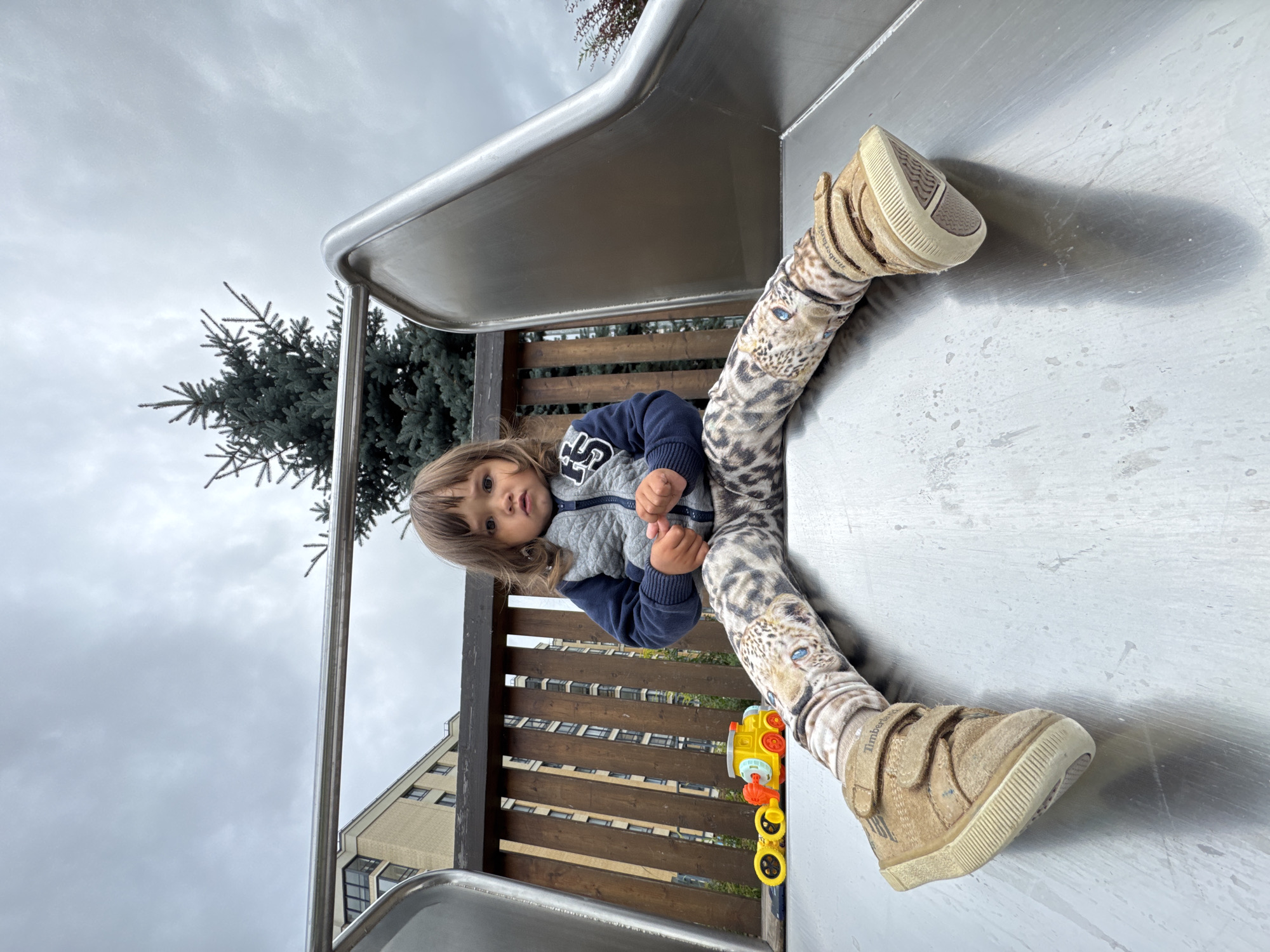 A young child with brown hair sits on a metal slide in a playground, looking directly at the camera. The child wears a grey and blue jacket, and patterned leggings with a leopard print. On the slide to the left of the child is a yellow and red toy train. The background shows a wooden fence, a tall evergreen tree, and a modern apartment building. The sky is overcast. The scene is located in the city of Novoselye, Russia.