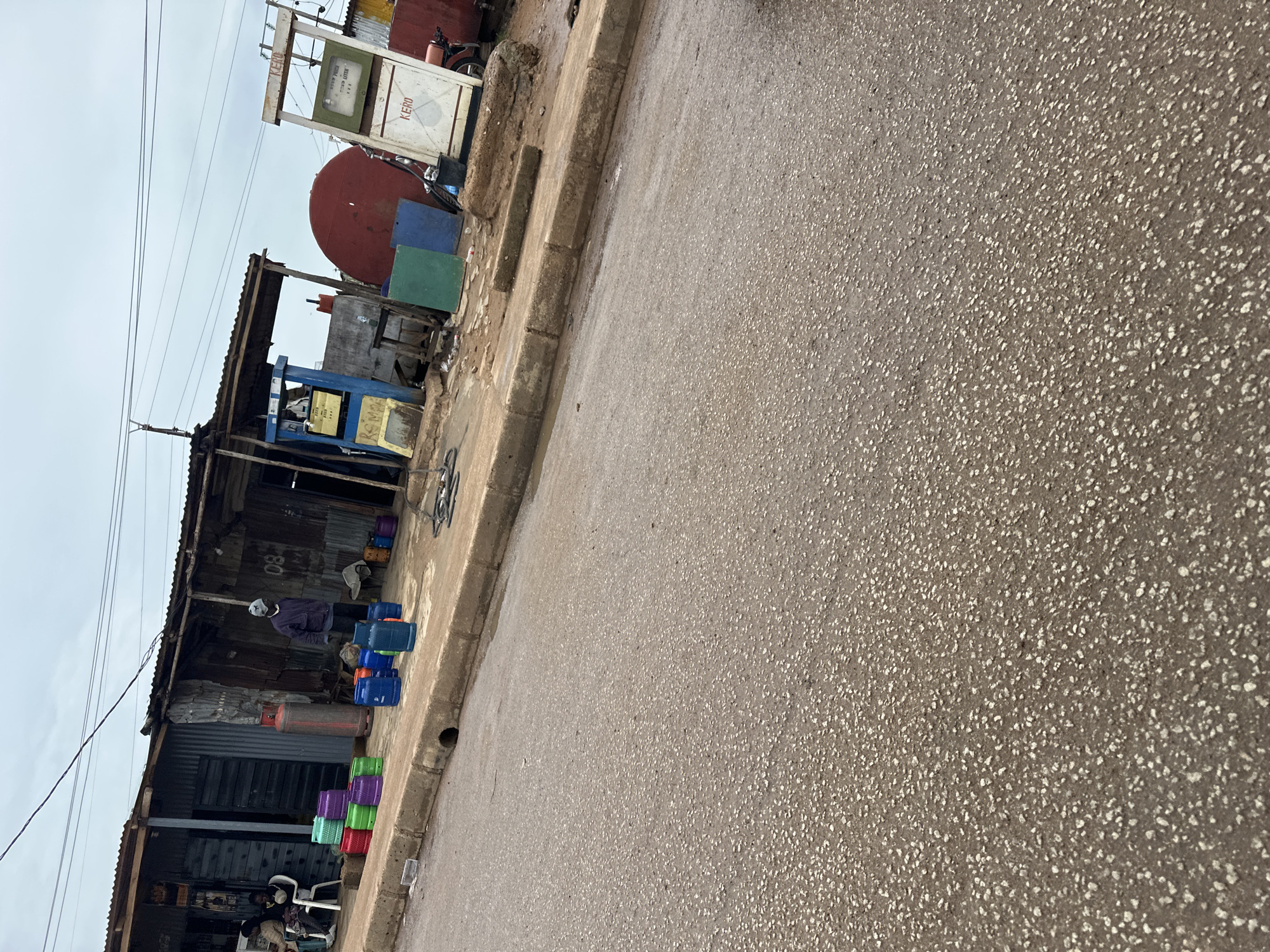 The image shows a street scene in Bukuru, Nigeria. In the foreground, there is a paved road. The middle ground features a sidewalk with various items on it, including colorful plastic containers, gas cylinders, and what appear to be two old-fashioned gasoline pumps. A person wearing a grey beanie and dark jacket stands near the gas pumps. Behind the sidewalk, there is a row of structures, likely shops, made of corrugated metal and wood. A large red tank is visible behind one of the structures. In the background, the sky is overcast. Two people are seated on chairs in front of one of the shops.