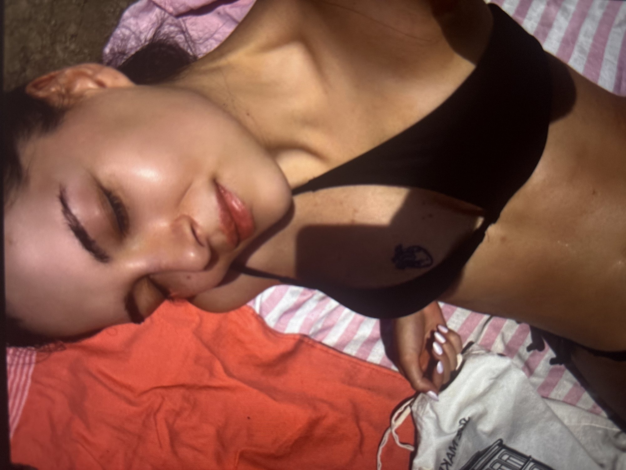 This media file depicts a young woman lying on a beach towel, seemingly sunbathing. She is wearing a black bikini top. Her eyes are closed, and her head is tilted back, suggesting relaxation. A small tattoo is visible on her upper chest. The setting appears to be outdoors, likely during the daytime, given the presence of sunlight casting shadows. The beach towel has a pink and white striped pattern, and an orange towel is also present. A white cloth bag with some text and an image is partially visible in the lower right corner. The woman's skin appears tanned, and she has light-colored nail polish on her fingers. The overall mood conveyed is one of leisure and enjoyment of the sun.