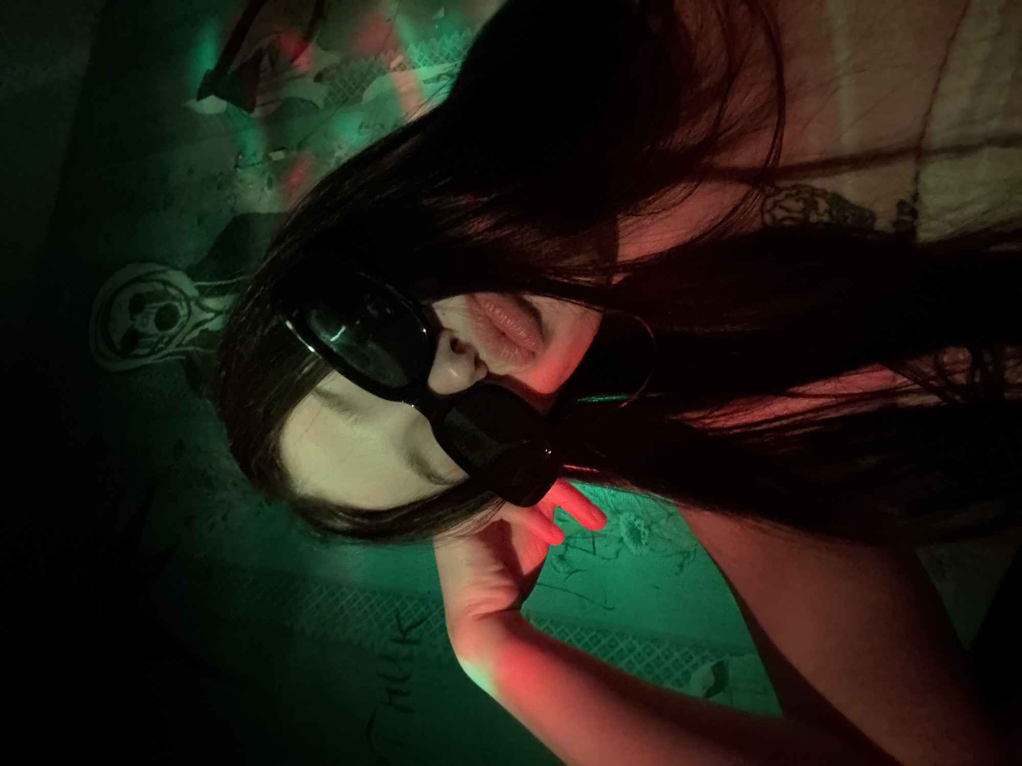 A person with dark hair and sunglasses is lying down on a patterned surface with green and red lighting. The person's right arm is visible. A drawing of a skull and the word "HULK" are on the patterned surface. The location cannot be determined from the image.